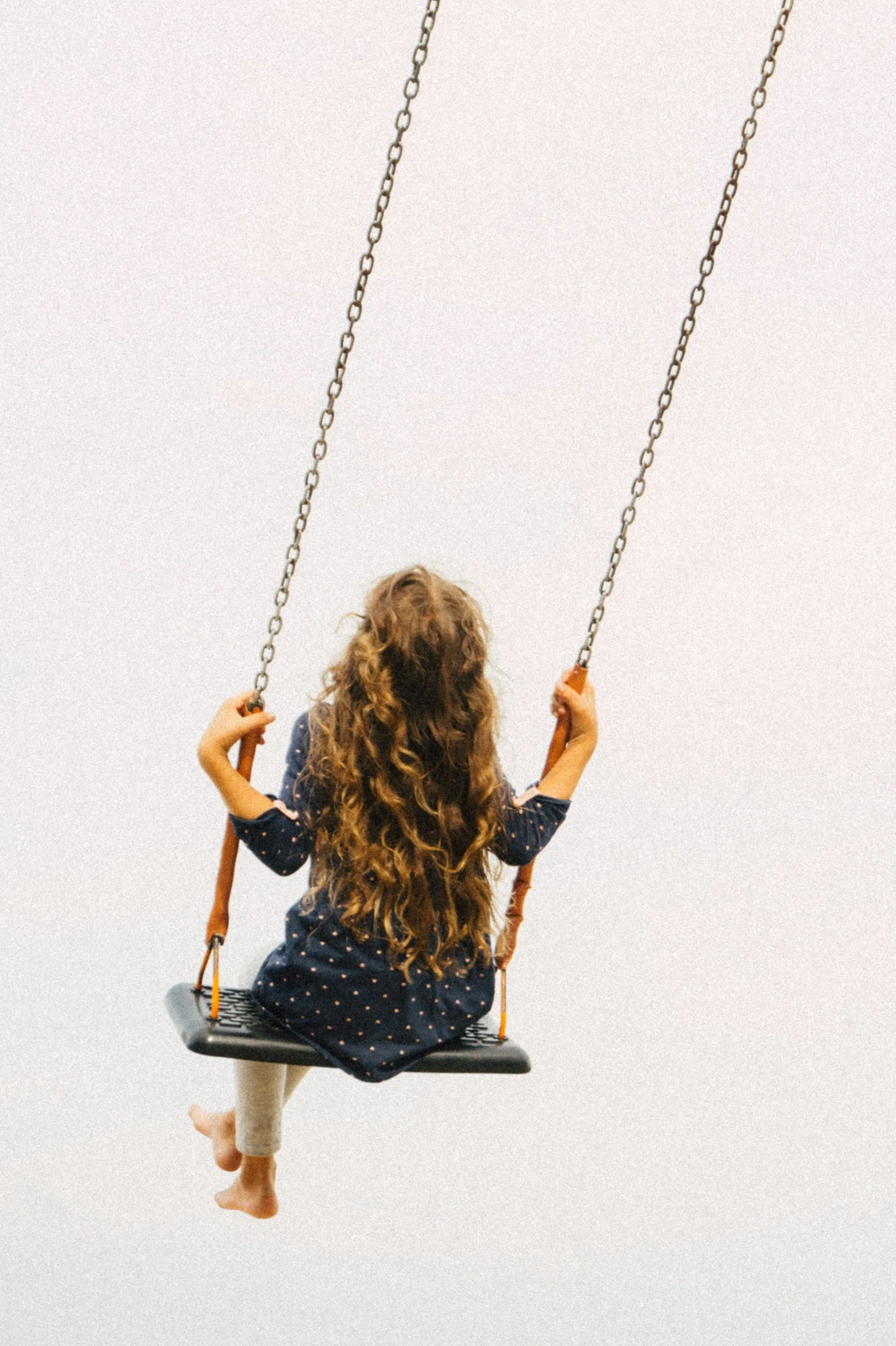 A young girl with long curly hair is sitting on a swing with a black seat and orange ropes, appearing to be swinging indoors against a plain, light-colored wall.