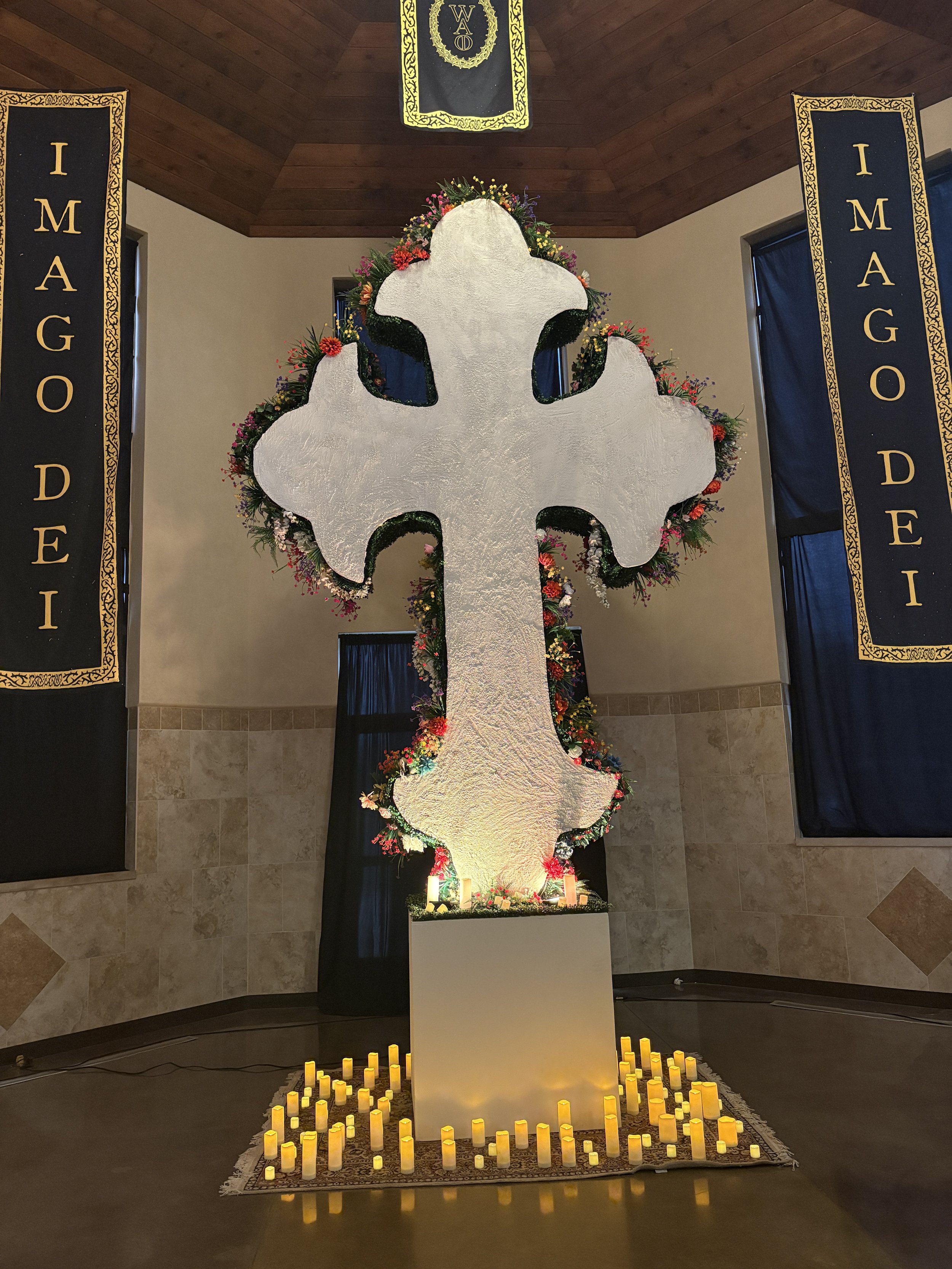 A white wood cross adorned with flowers in pink, purple, orange, and green. Surrounding the base of the cross are numerous lit yellow candles, placed on a decorative rug. The background features two black banners with gold lettering and gold borders, reading 'IMAGO DEI,' and a black banner hanging from the ceiling with a gold design and the abbreviation 'WAD'.