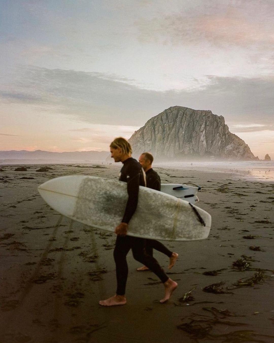 Two surfers walking on a beach carrying surfboards, with a large rock formation in the background during sunset.