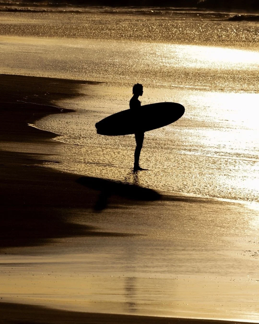Silhouette of a person carrying a surfboard, standing on the beach at sunset with reflections on the wet sand and gentle waves.