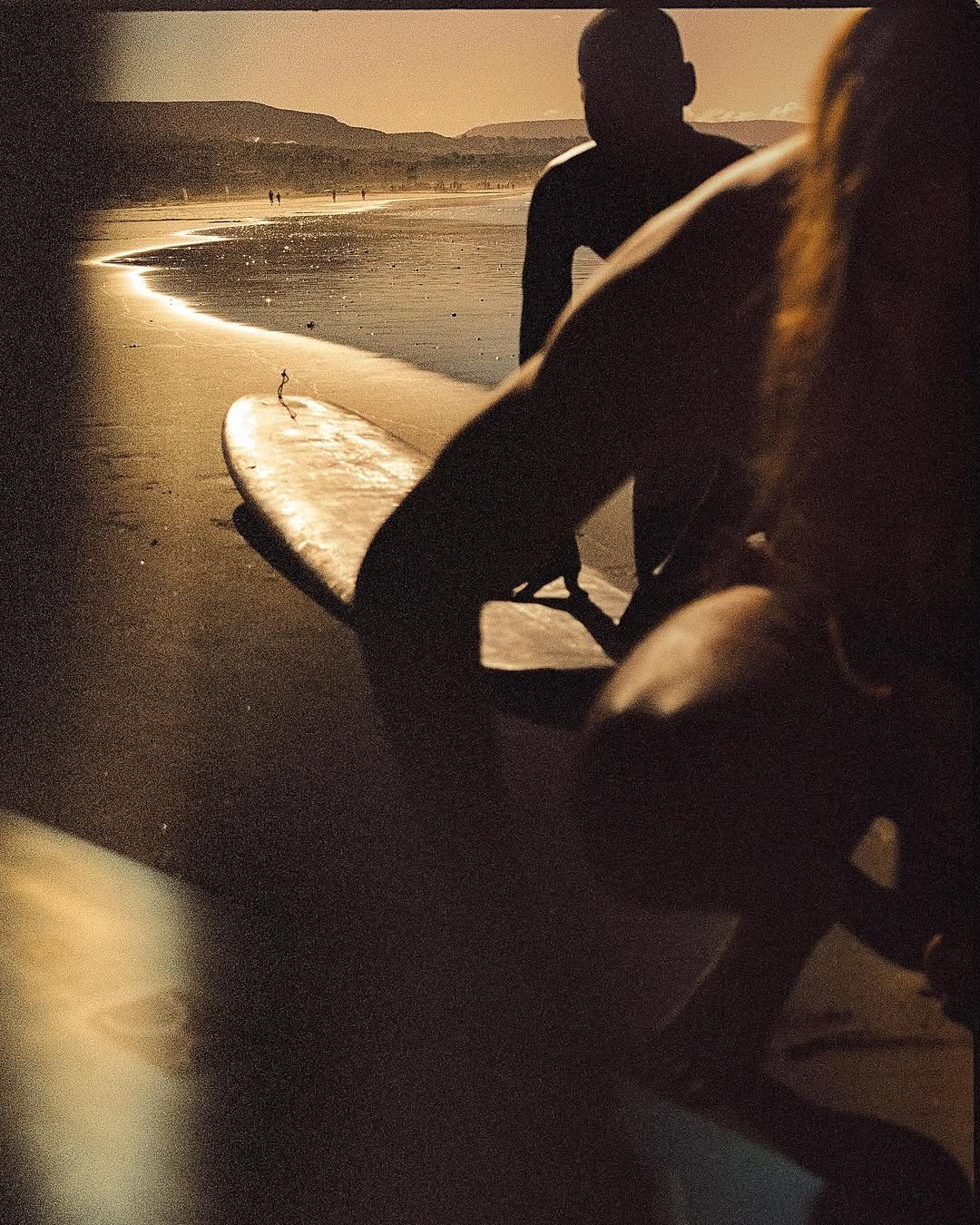 Silhouettes of two people on a beach at sunset, with one holding a surfboard and distant people walking along the shoreline.