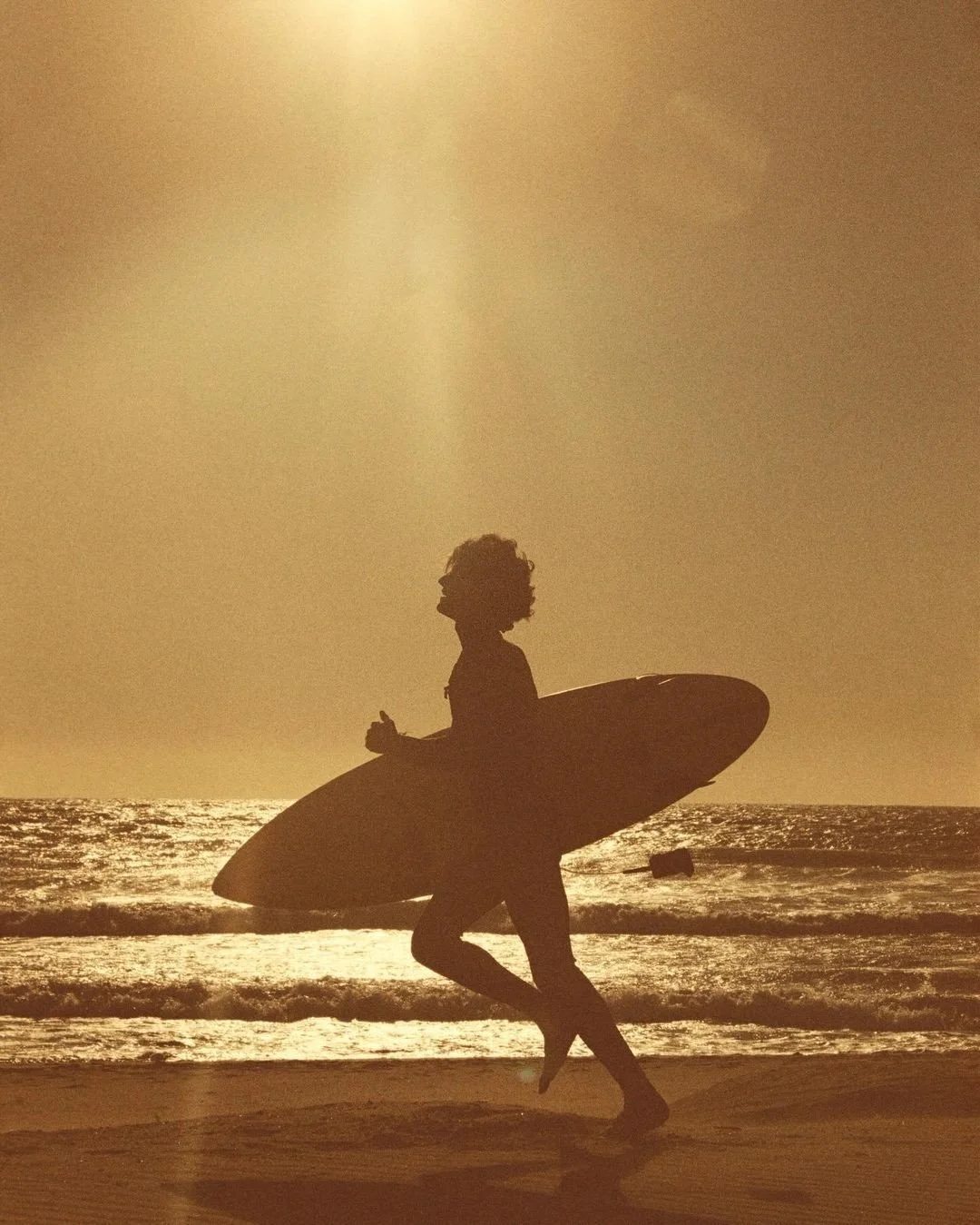 A person holding a surfboard walking on the beach during sunset or sunrise, with the ocean and sky in the background.