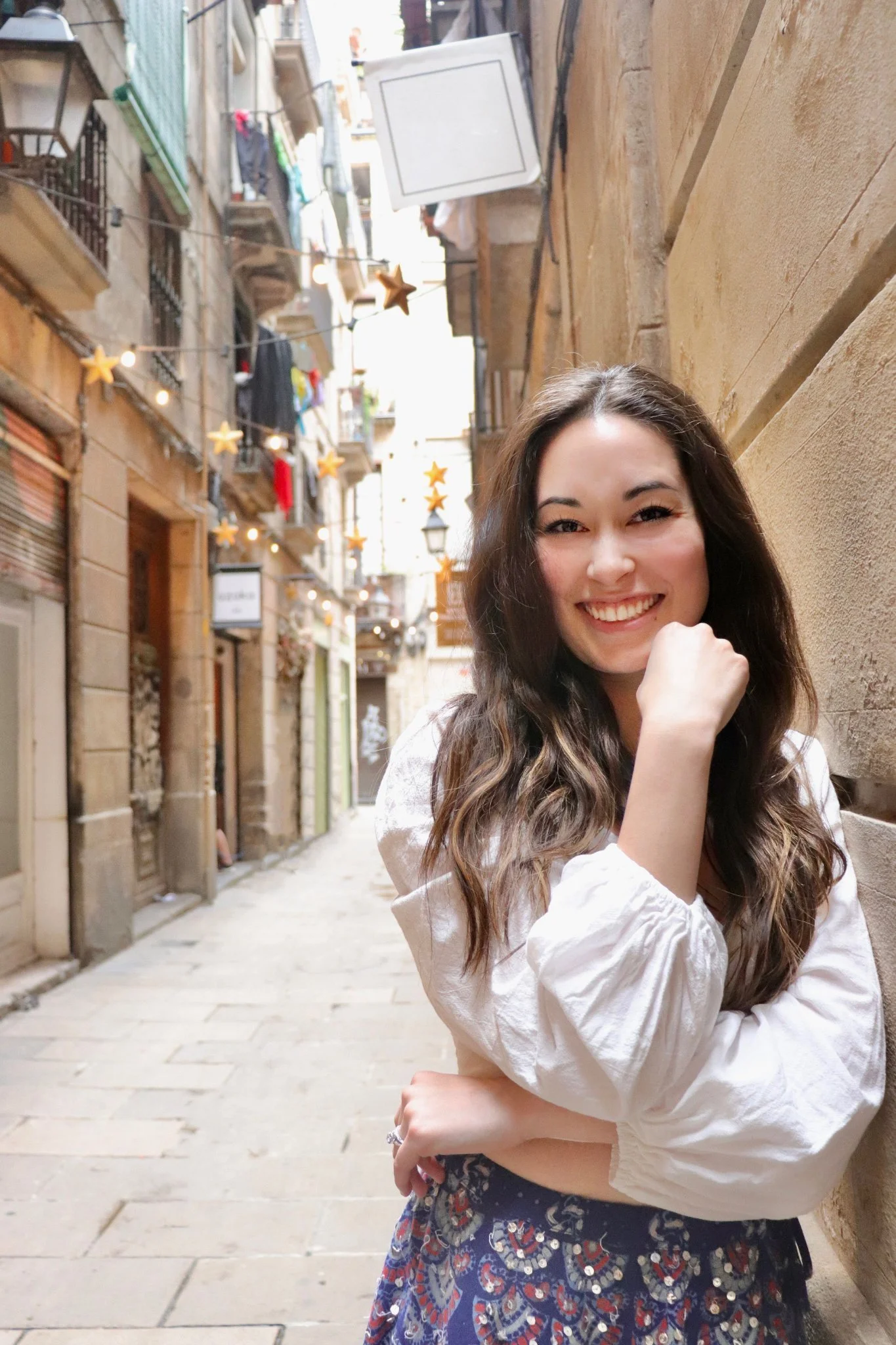 A smiling young woman with long brown hair stands in a narrow street with European-style buildings. She is wearing a white blouse with puffed sleeves and a blue skirt with decorative patterns. The street is decorated with hanging string lights and star-shaped ornaments.