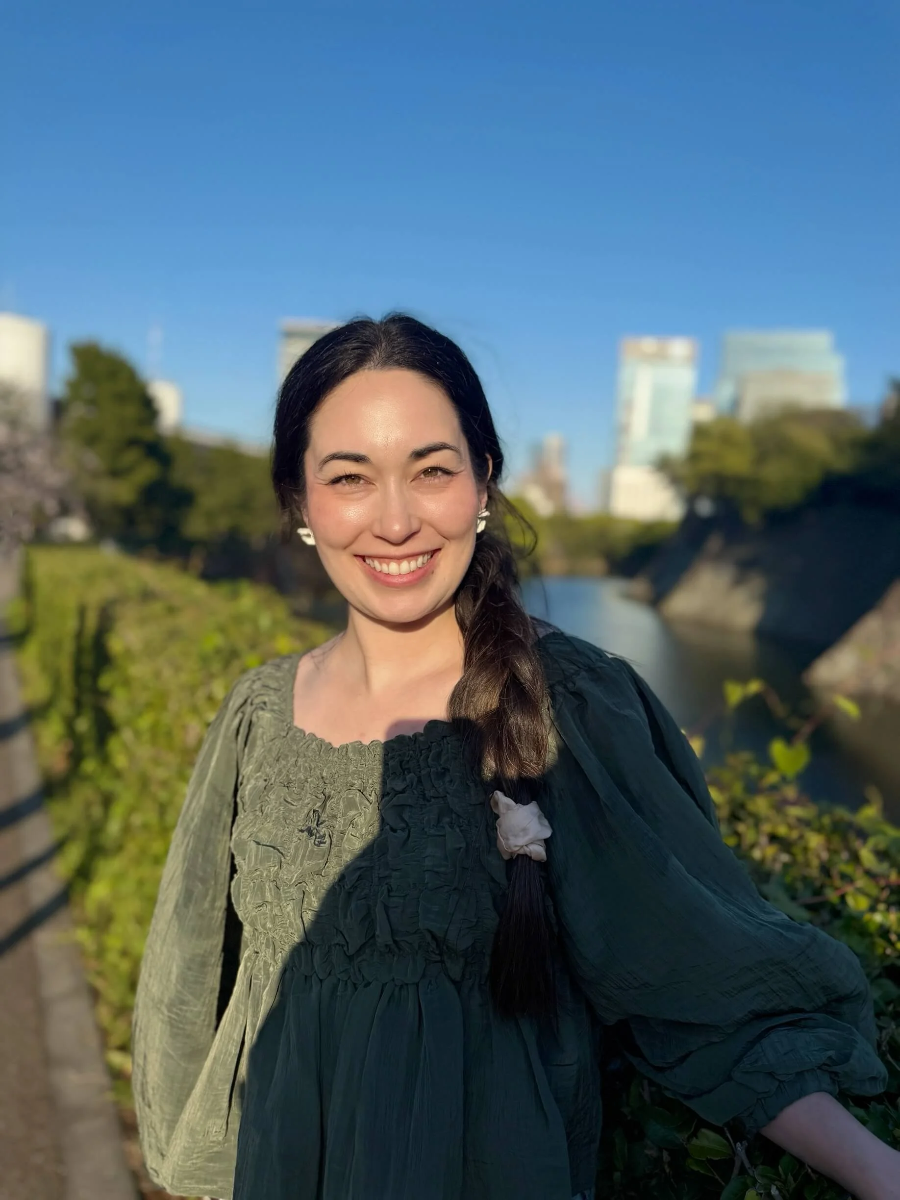 Smiling woman with dark hair in a braid, wearing earrings and a dark jacket, standing outdoors near a river and city buildings under a clear blue sky.