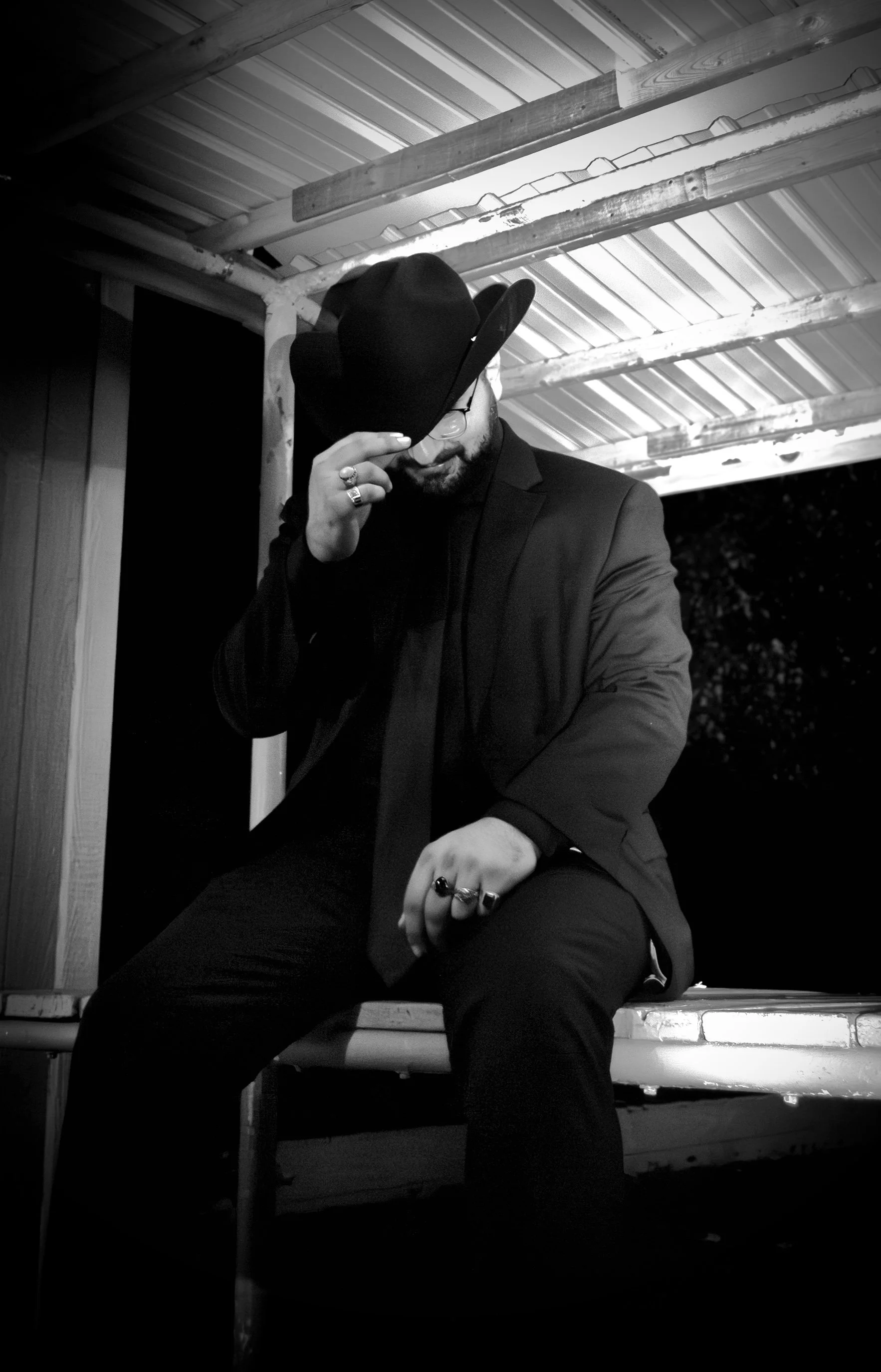 Black and white photo of a man wearing a suit, hat, and glasses, sitting on a bench under a metal roof, adjusting his hat with one hand and resting his other hand on his knee with rings, during nighttime.