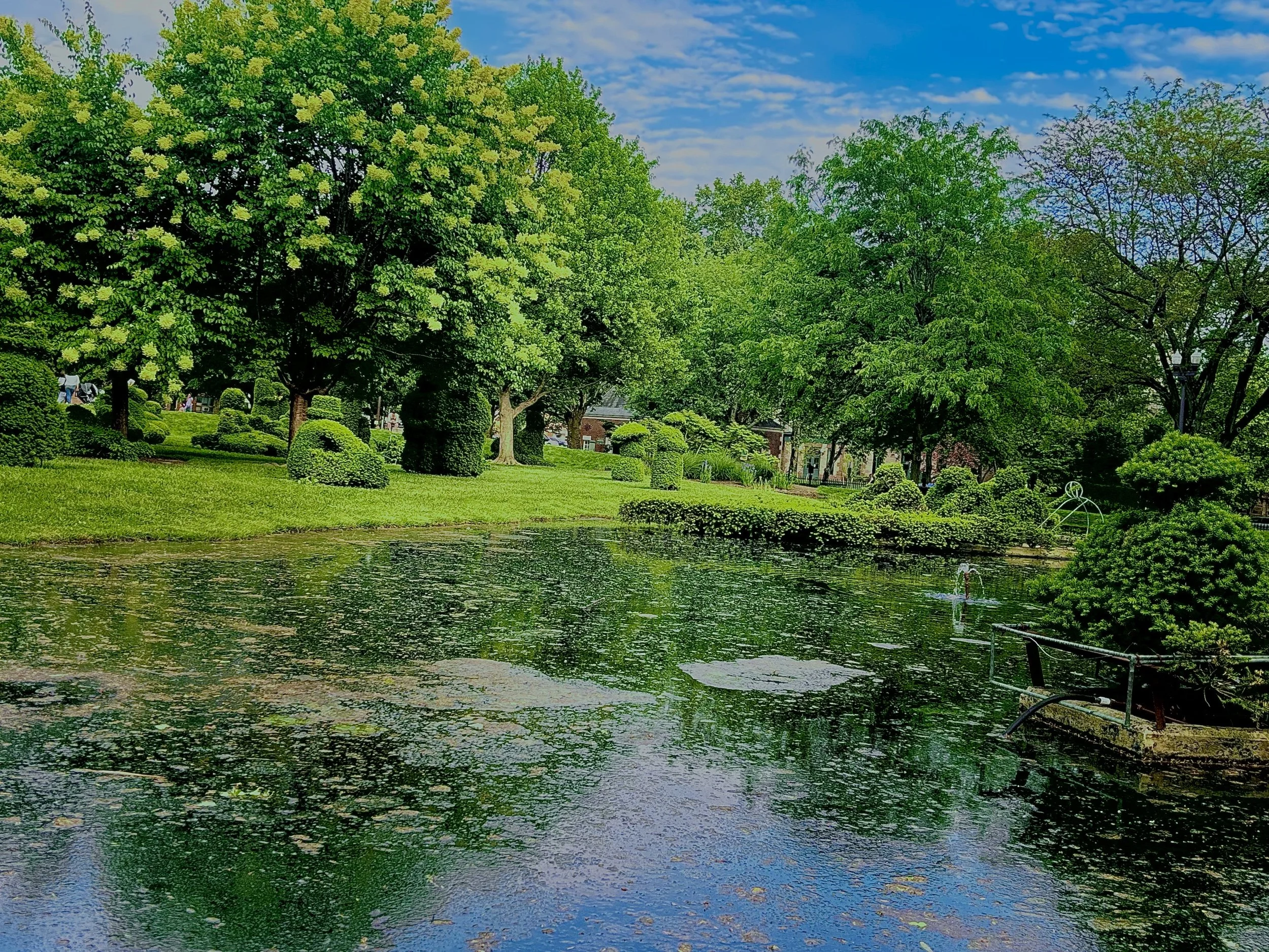 A park scene with lush green trees, well-manicured bushes, and a pond with lily pads and reeds. There are people in the background near houses or buildings, and the sky is partly cloudy with blue patches.