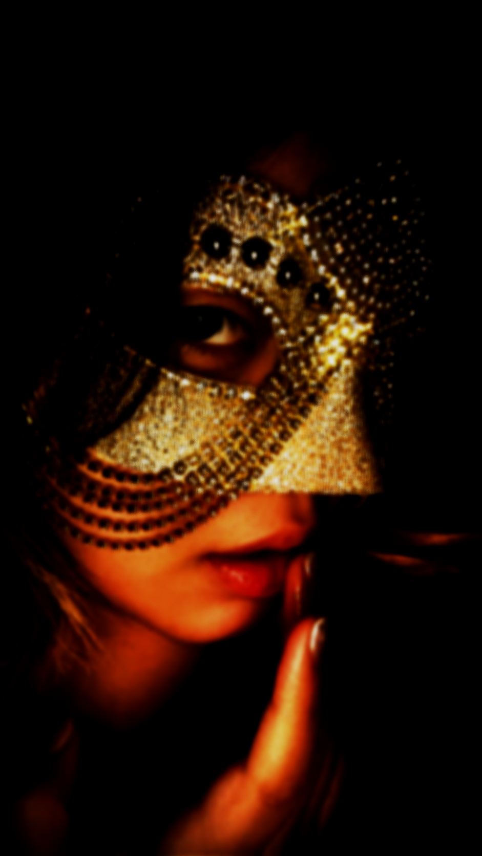 A woman wearing an ornate, rhinestone-encrusted mask covering the upper half of her face, with her finger to her lips in a shushing gesture, against a dark background.