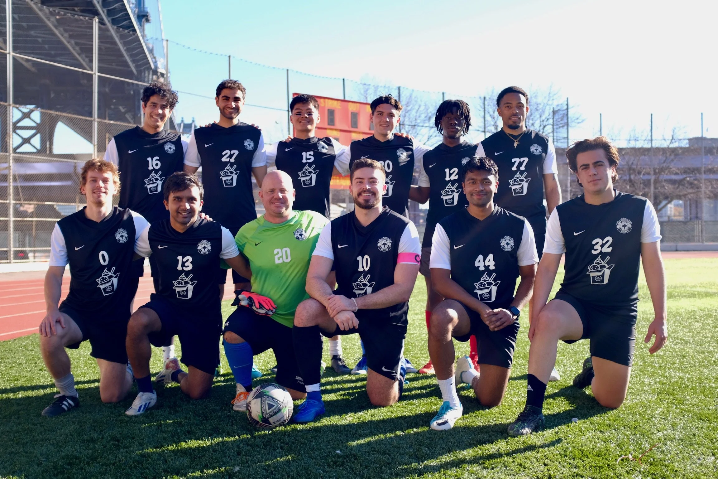 A group of ten male soccer players and their coach posing for a team photo outdoors on a soccer field, with a bright blue sky and stadium seating in the background.