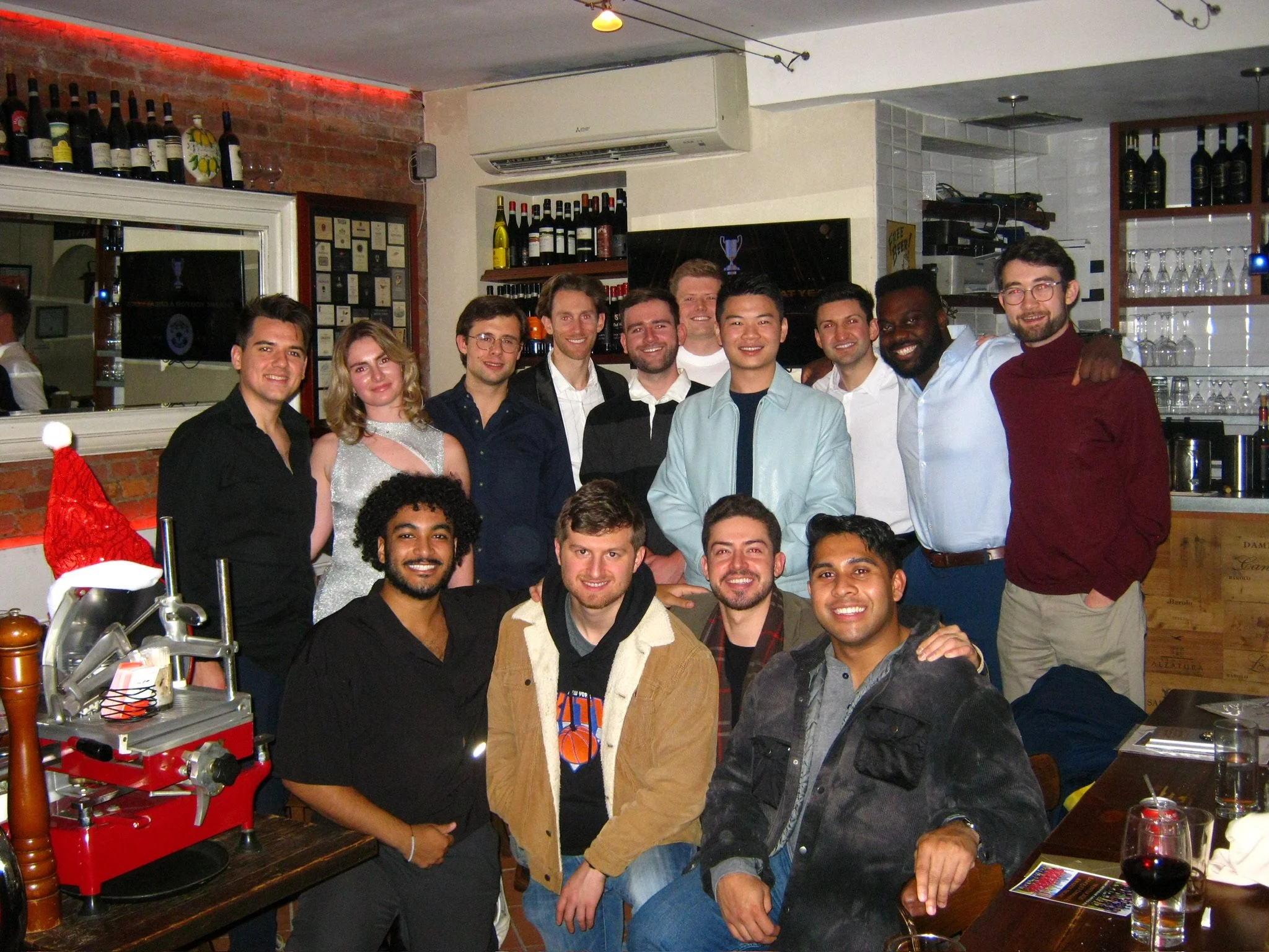 A group of diverse young adults gathered together in a restaurant, smiling at the camera, with wine bottles and a bar in the background.
