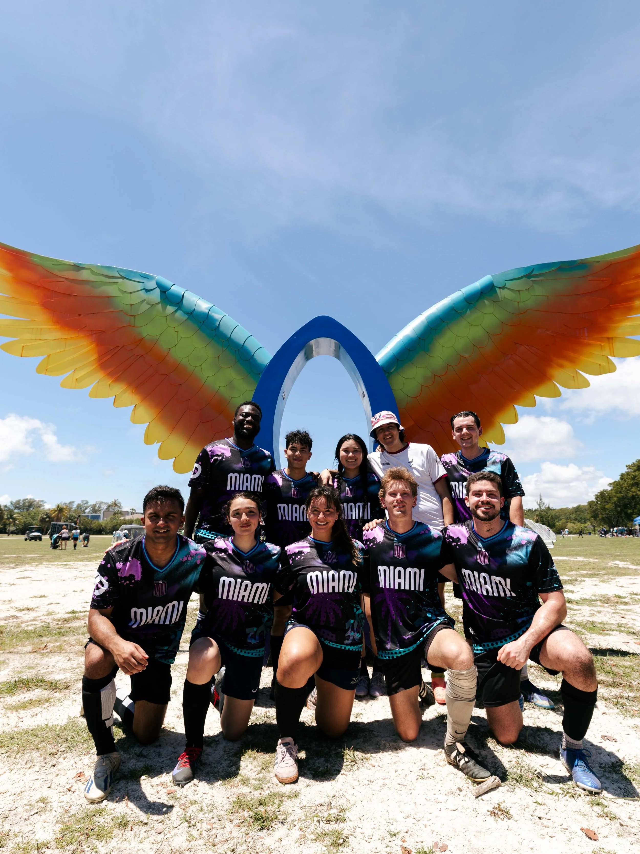 A group of young people wearing Miami sports uniforms posing outdoors in front of a large decorative rainbow-colored wing sculpture during the day.