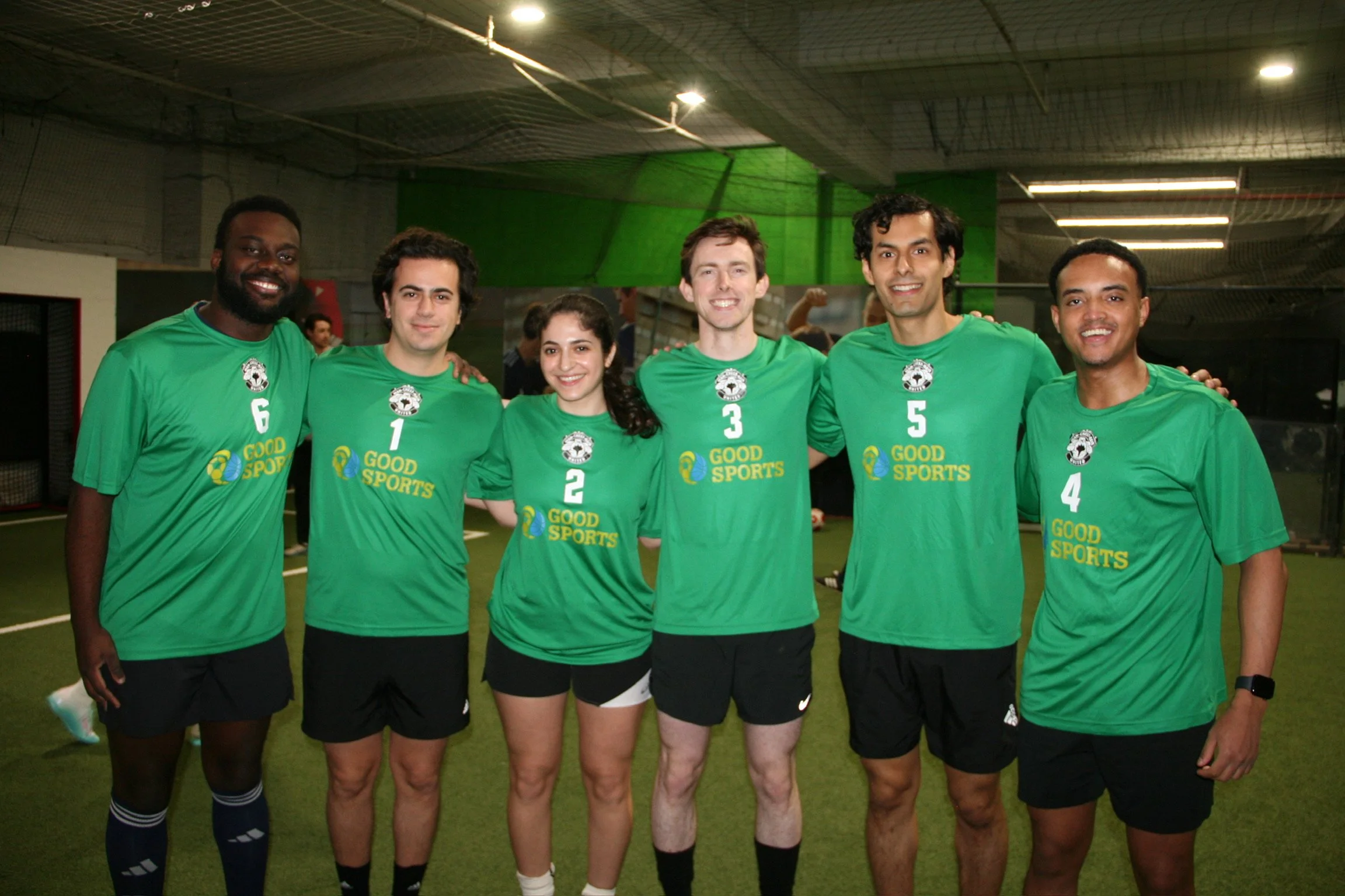 A group of six young adults, three men and three women, in green soccer jerseys with numbers 6, 1, 2, 3, 5, and 4, smiling and posing together inside an indoor sports facility.