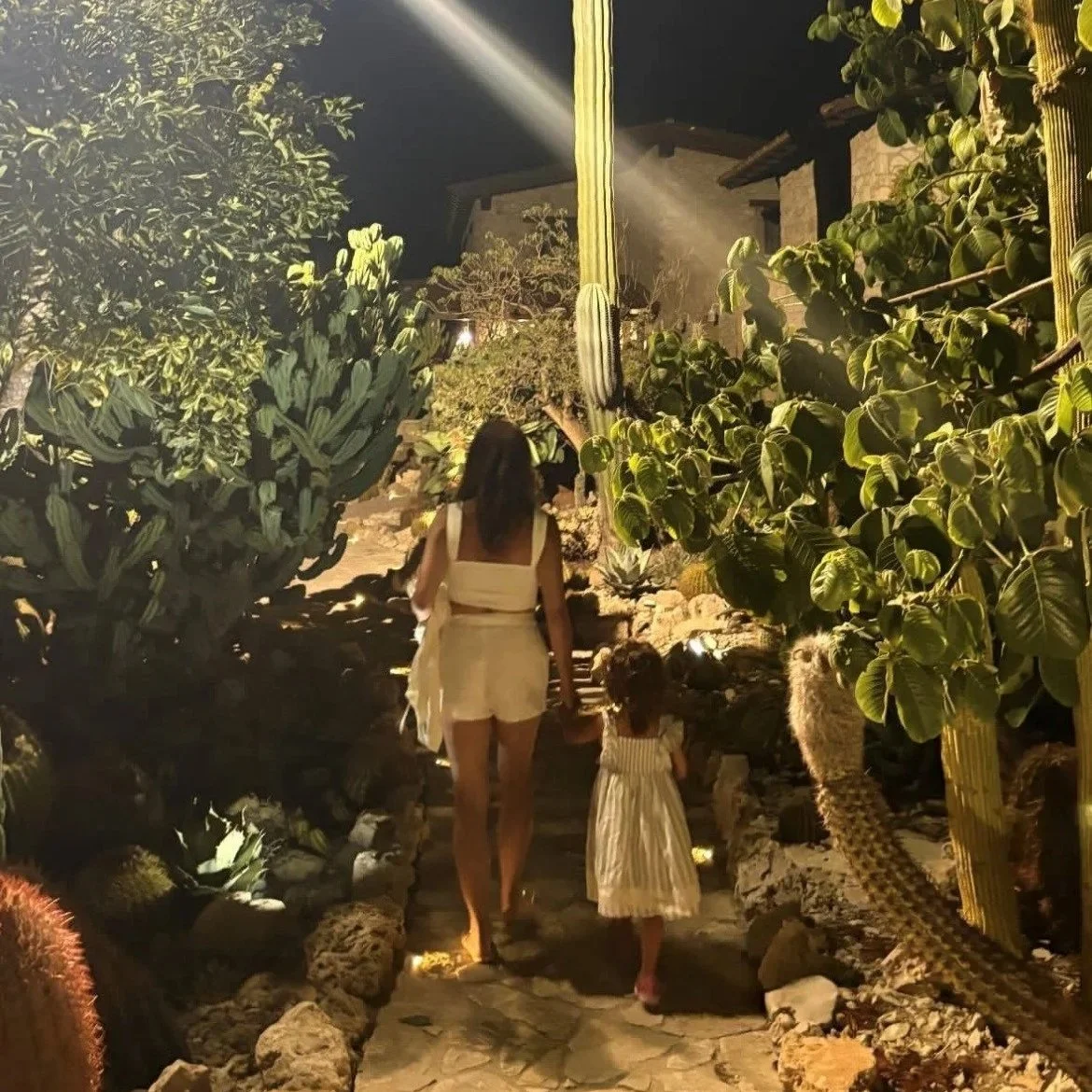 A woman and young girl walk hand in hand along a stone pathway at night. The pathway is surrounded by desert plants, including cacti and succulents, illuminated by outdoor lighting.