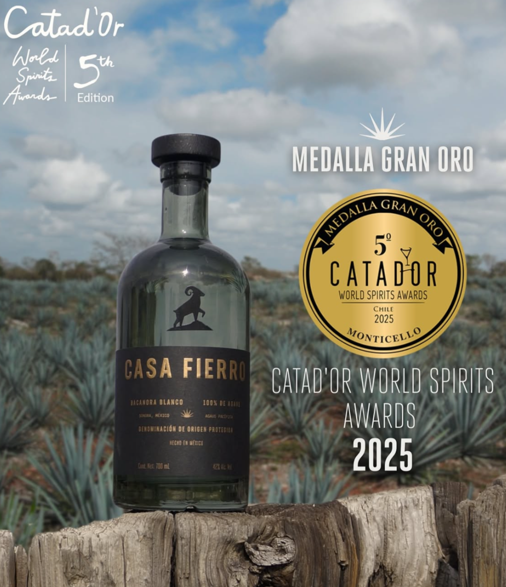 Bottle Casa Fierro Bacanora Blanco tequila mezcal on wooden surface with an agave field farm in the background. The image features a Gran Oro Grand Gold medal it received at 2025 Catad'Or World Spirits Awards, held in Monticello, Chile.