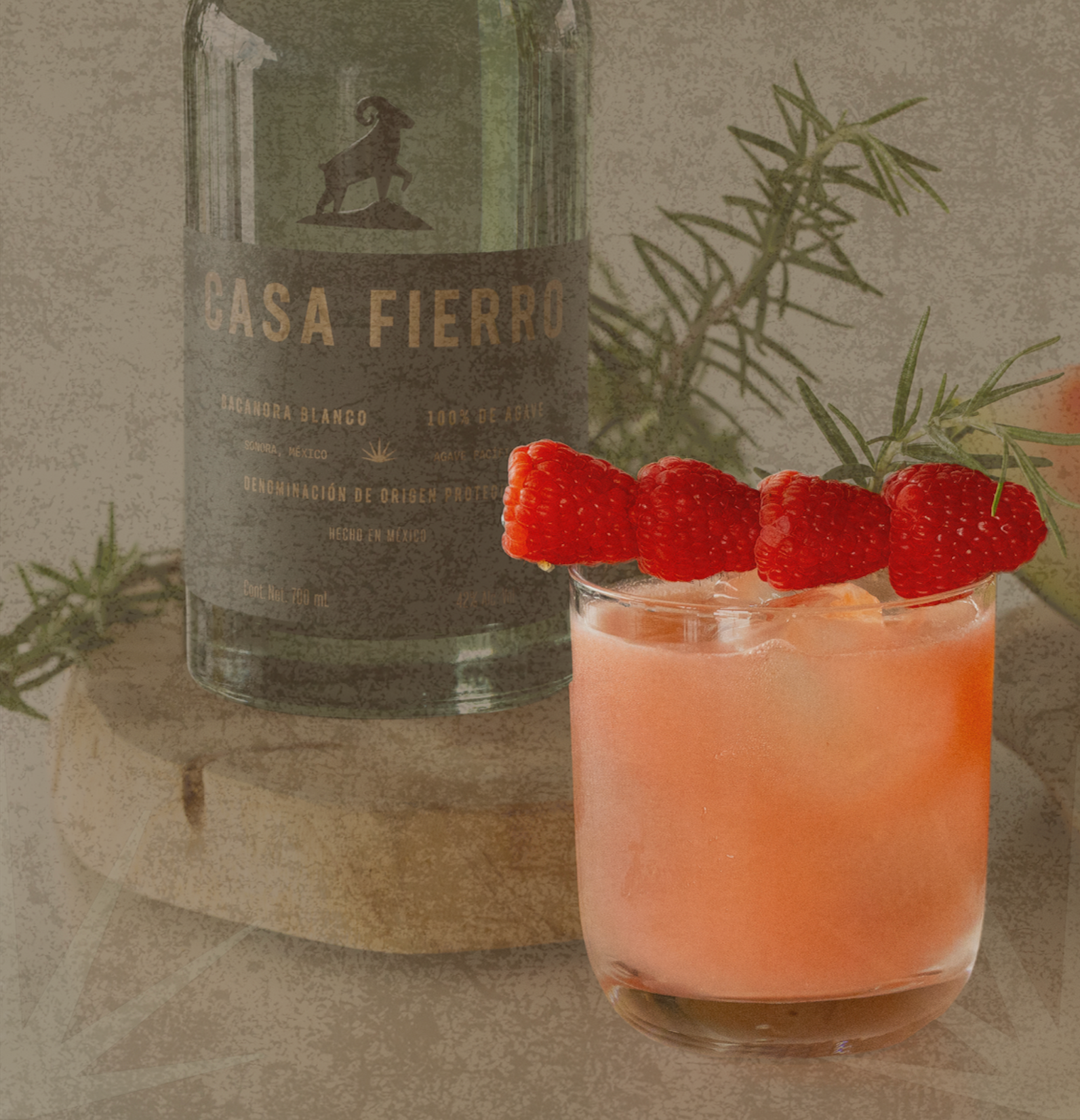 A glass of pinkish cocktail topped with fresh raspberries and garnished with a sprig of rosemary, placed in front of a bottle of Casa Fierro alcohol with a rosemary branch beside it on a wooden surface.