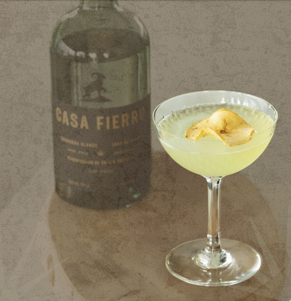 A cocktail glass with a pale yellow drink and apple slices on top, placed on a wooden surface. In the background, there is a dark bottle with the label 'Casa Fiero'.