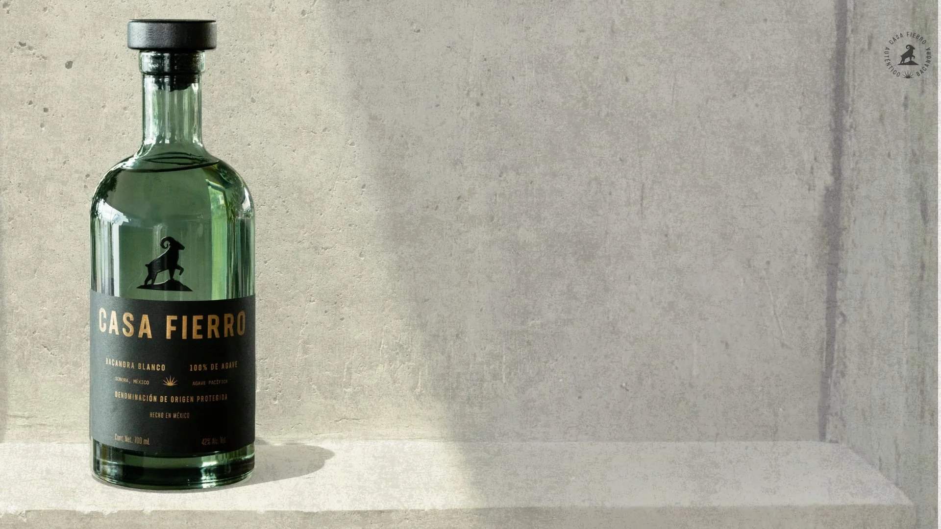 Dark green grey bottle Casa Fierro Agave Spirit Bacanora Blanco Tequila Mezcal black label and copper colored label standing before a light grey textured wall.