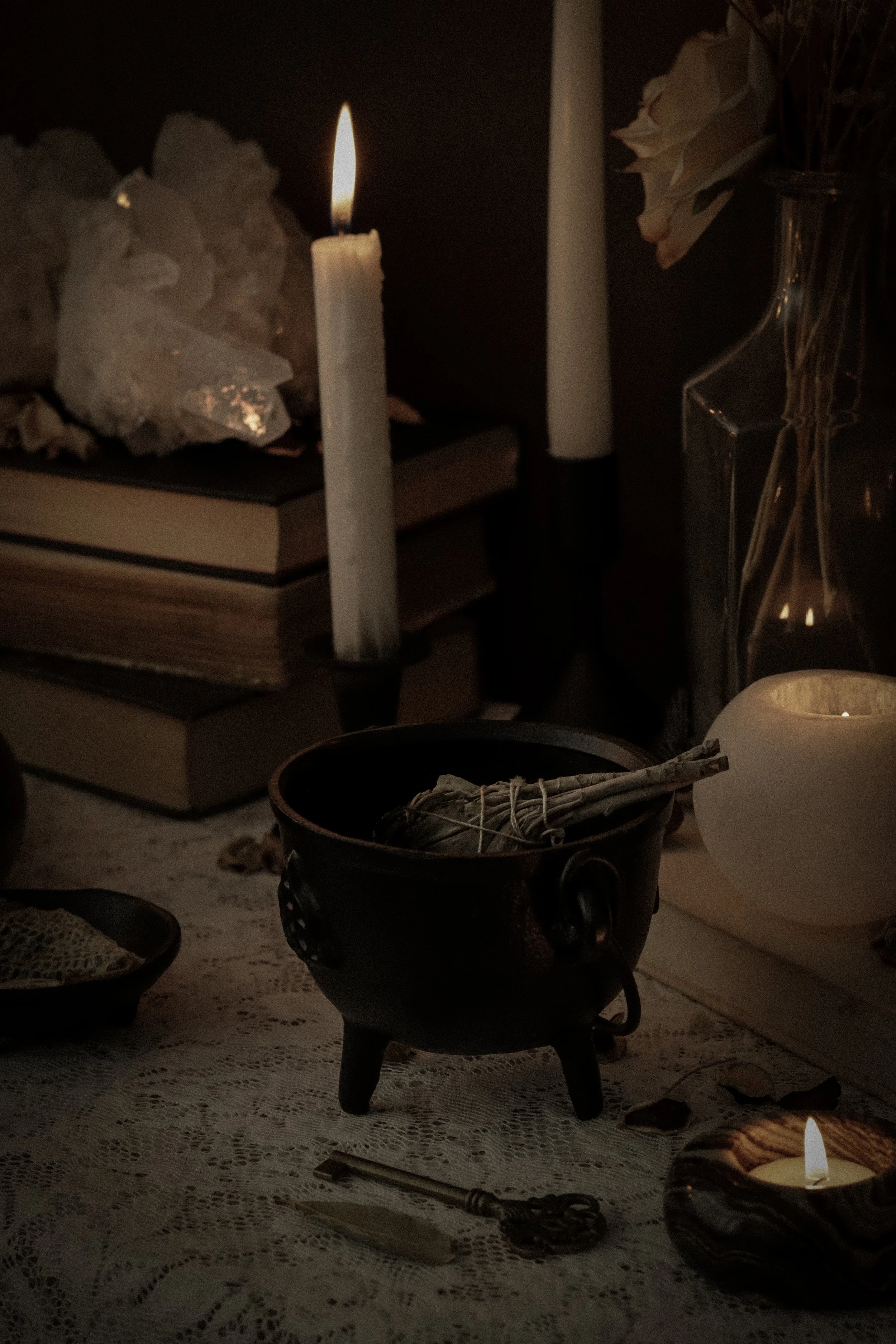 A dimly lit scene with a lit white candle, a book beneath rocks, a glass vase with dried roses, a black bowl with rolled paper, and a small candle on a lace tablecloth.