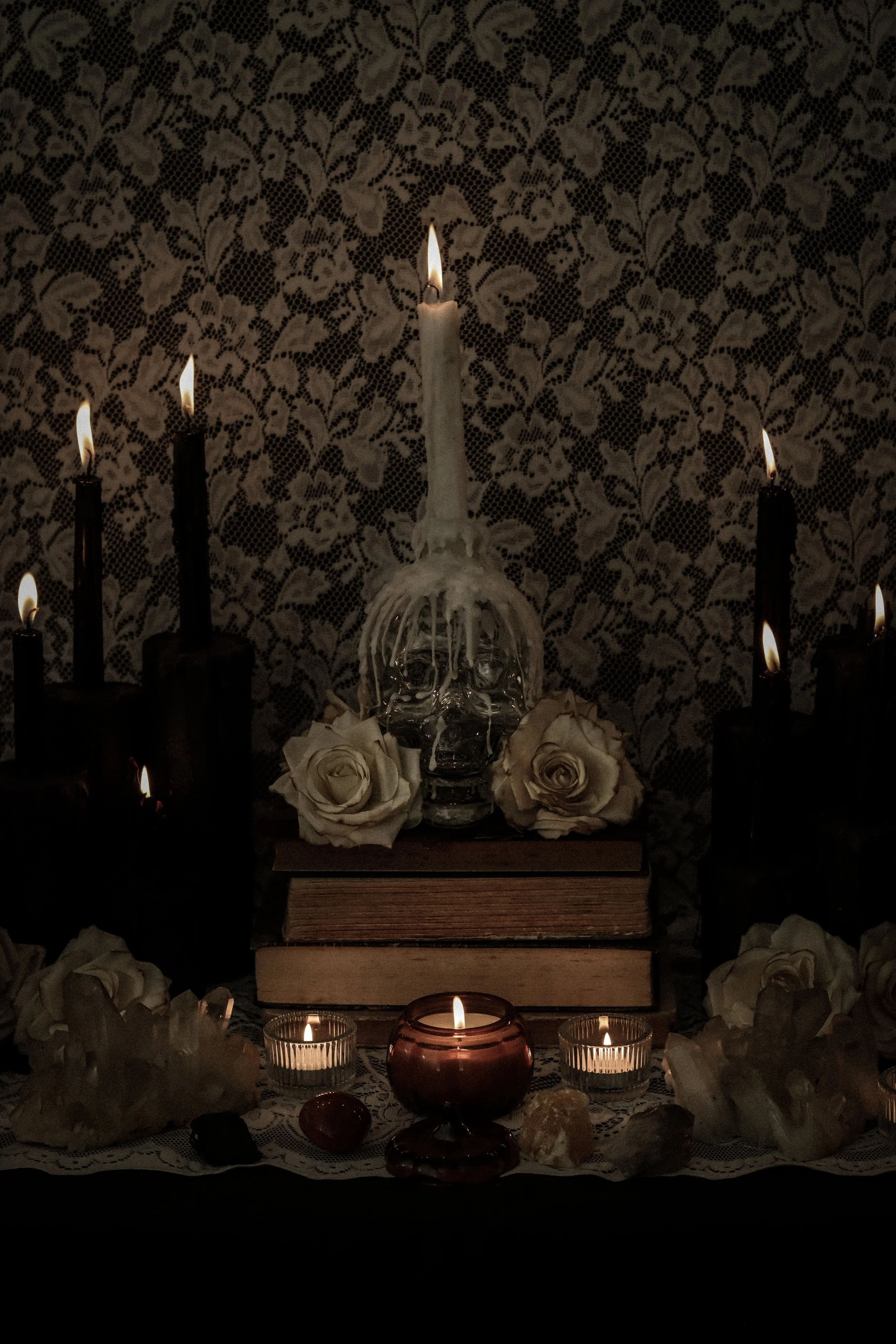 A vintage altar with lit white candles, white roses, and small candles in glass holders against a black and white lace floral patterned background.
