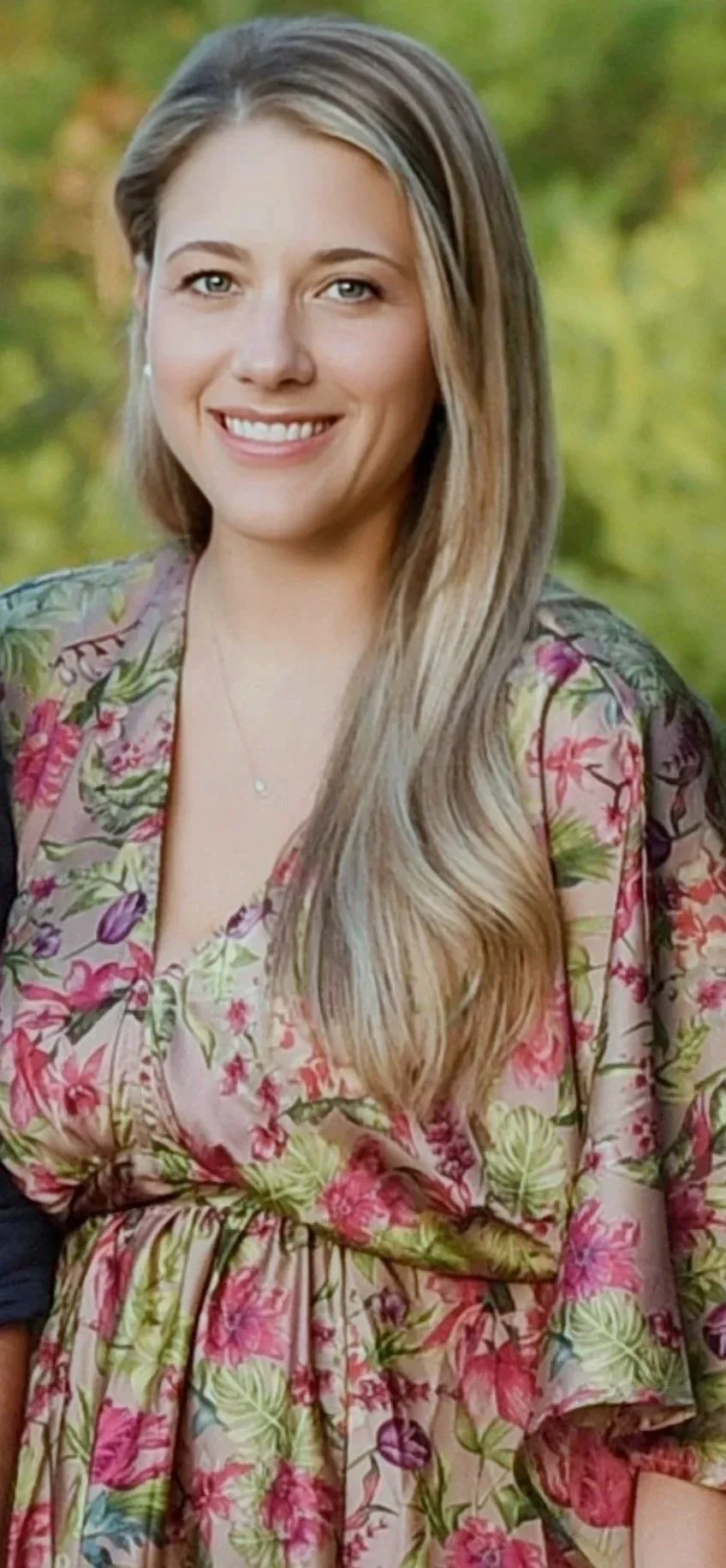 A young woman with long, wavy blonde hair, green eyes, and a bright smile, wearing a floral dress with pink, purple, and green patterns, standing outdoors with green foliage in the background.