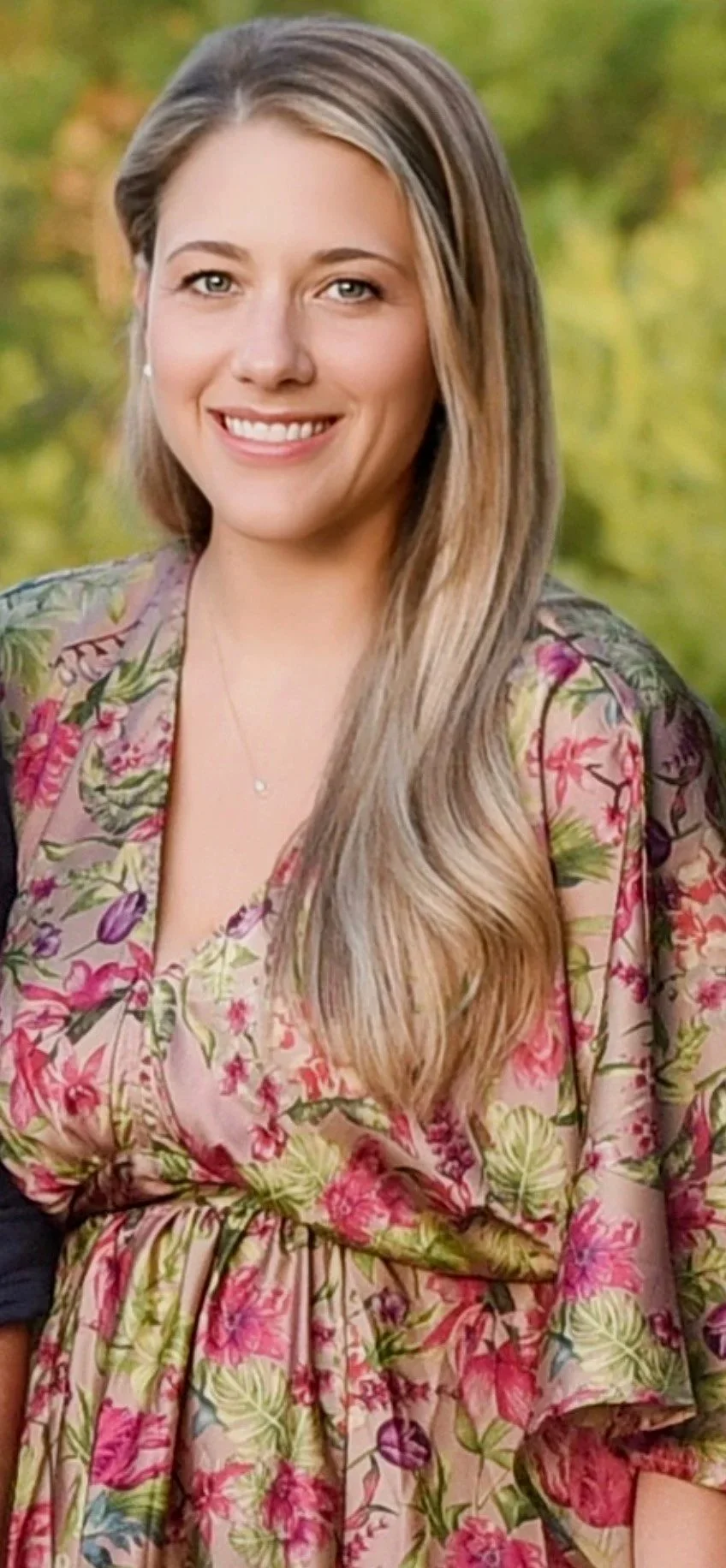 A smiling woman with long, wavy blonde hair, wearing a floral-patterned dress and standing outdoors with green trees in the background.