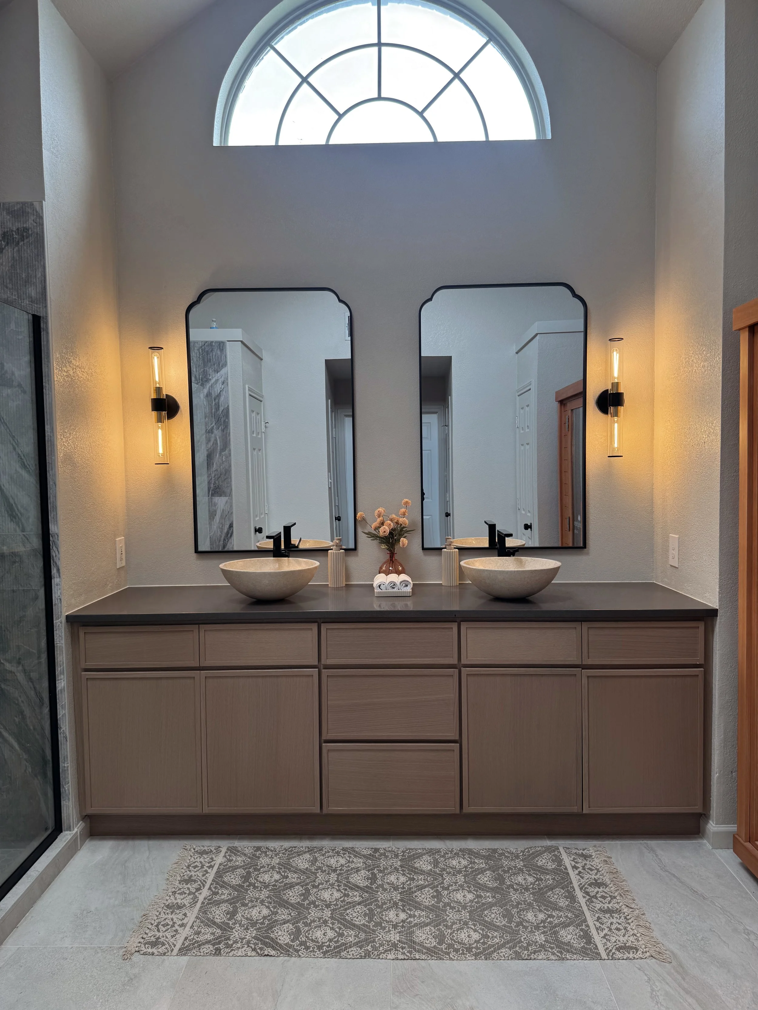 Modern bathroom vanity with two vessel sinks, black fixtures, large front mirrors, and decorative lighting. There is a flower arrangement and rolled towels between the sinks, with a patterned rug on the light-colored floor.