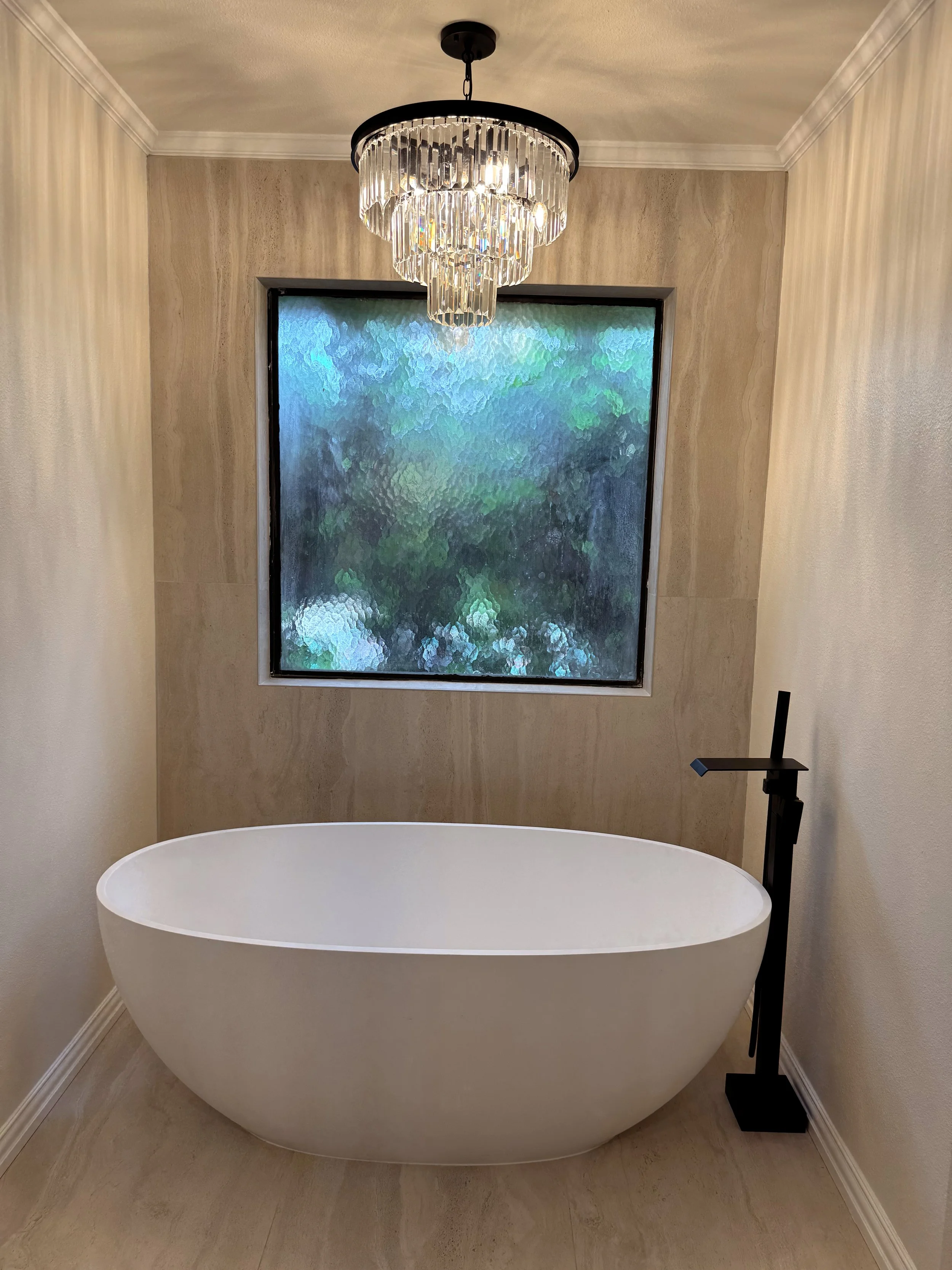 A modern freestanding bathtub in a bathroom with a large textured window and a black chandelier hanging from the ceiling.