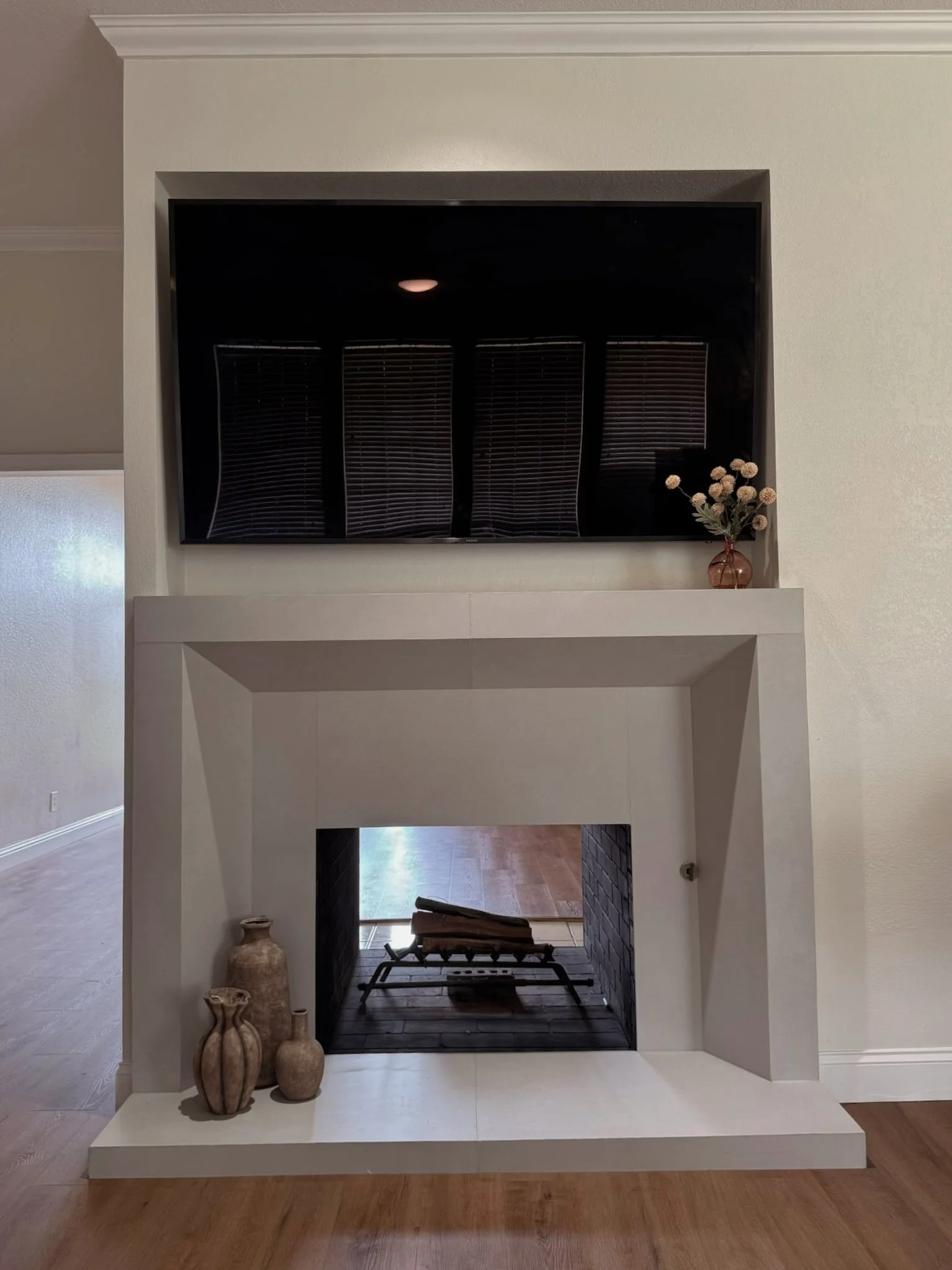 Interior view of a living room fireplace with a white mantel. Decor items include three beige vases on the left side of the mantel and a small brown vase with dried flowers on the right side. The fireplace contains a stack of logs and a fireplace gra
