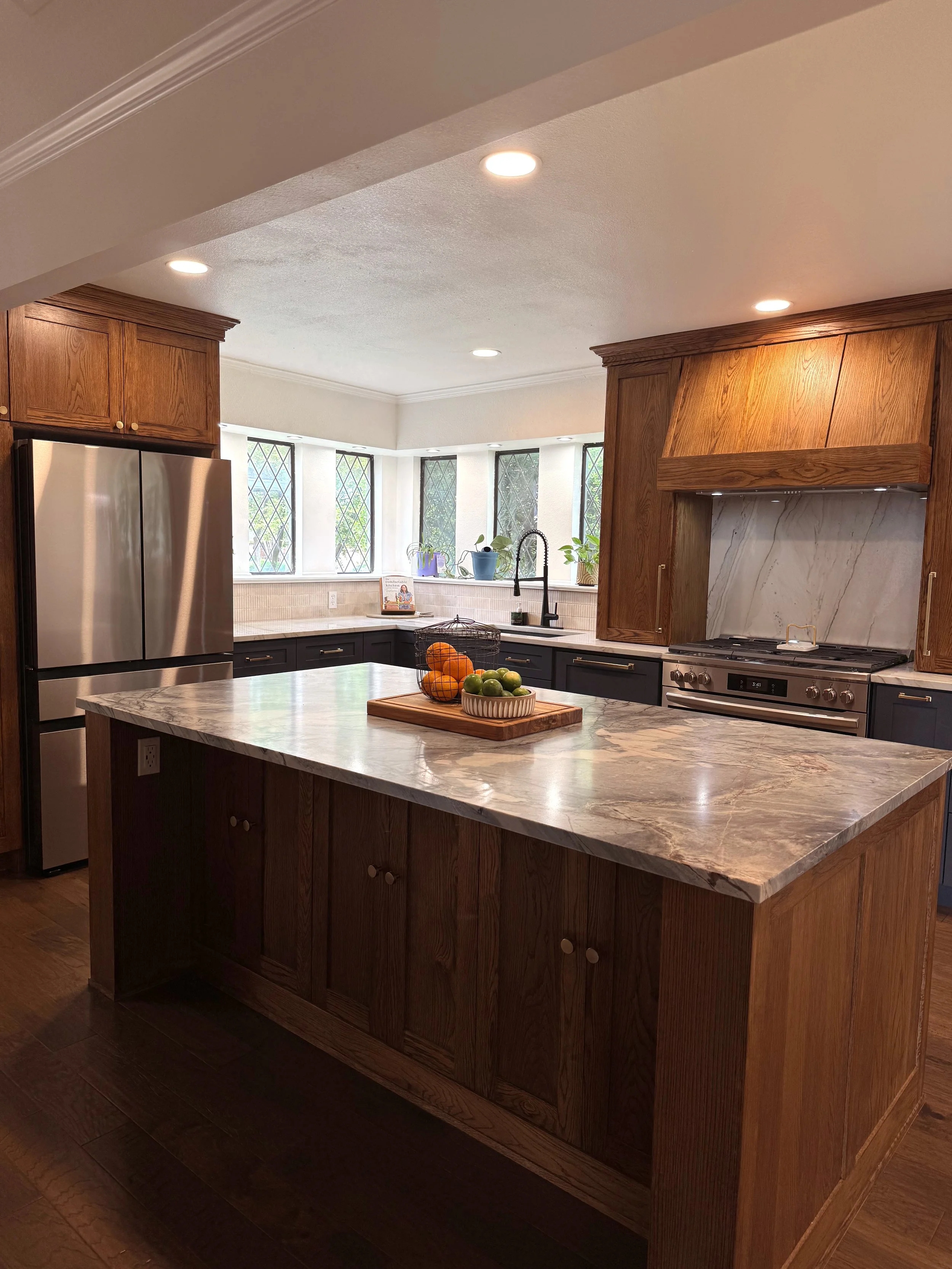 A contemporary kitchen with a marble countertop island, dark lower cabinets, and wooden upper cabinets. Stainless steel refrigerator and gas stove are visible, with a windowed backsplash and potted plants in the background.