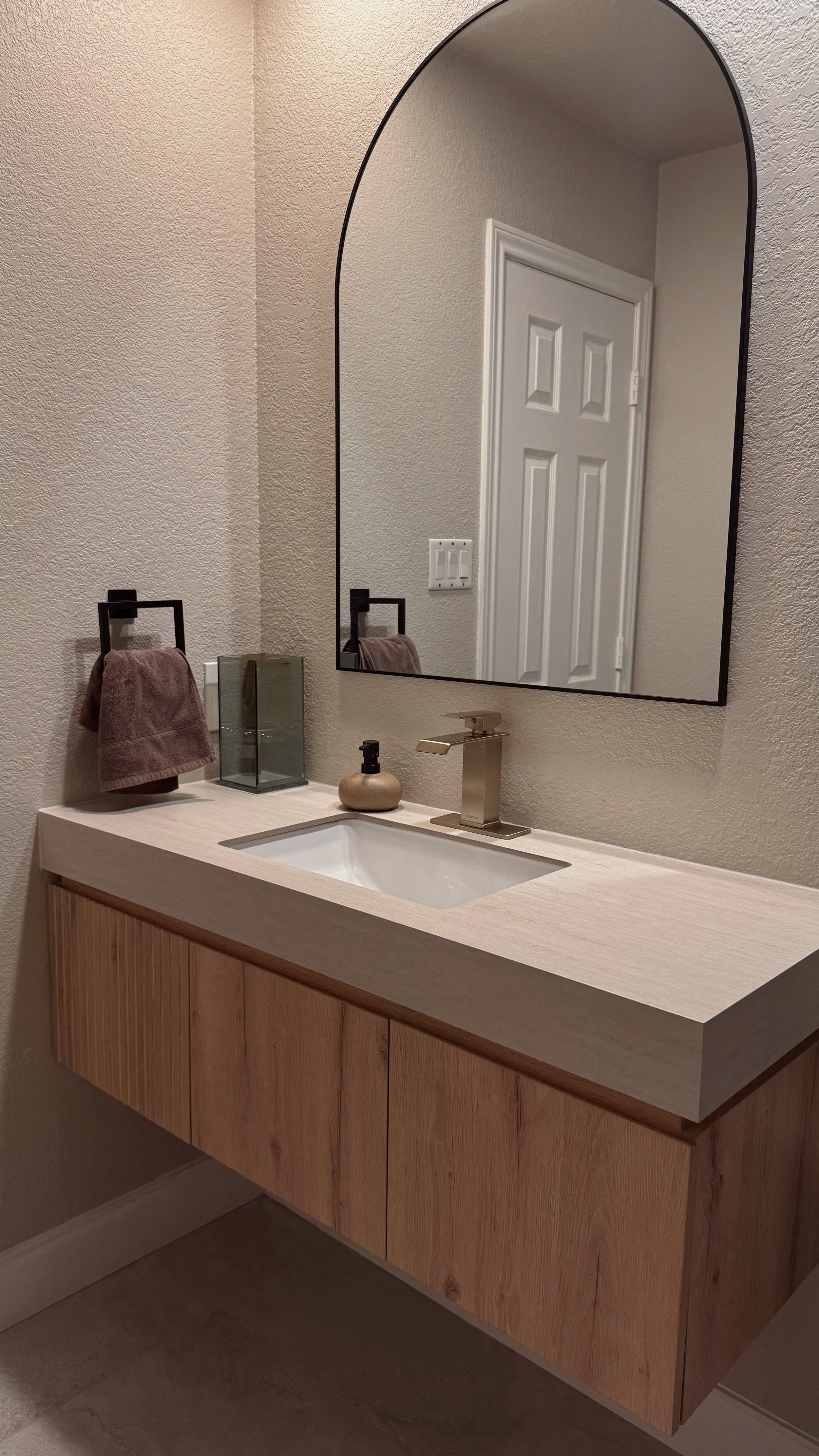 Bathroom vanity with a rectangular sink, gold faucet, a soap dispenser, a glass container, and a towel hanging on a black towel ring. Mirror on the wall above the vanity, door reflected in the mirror, beige textured walls.