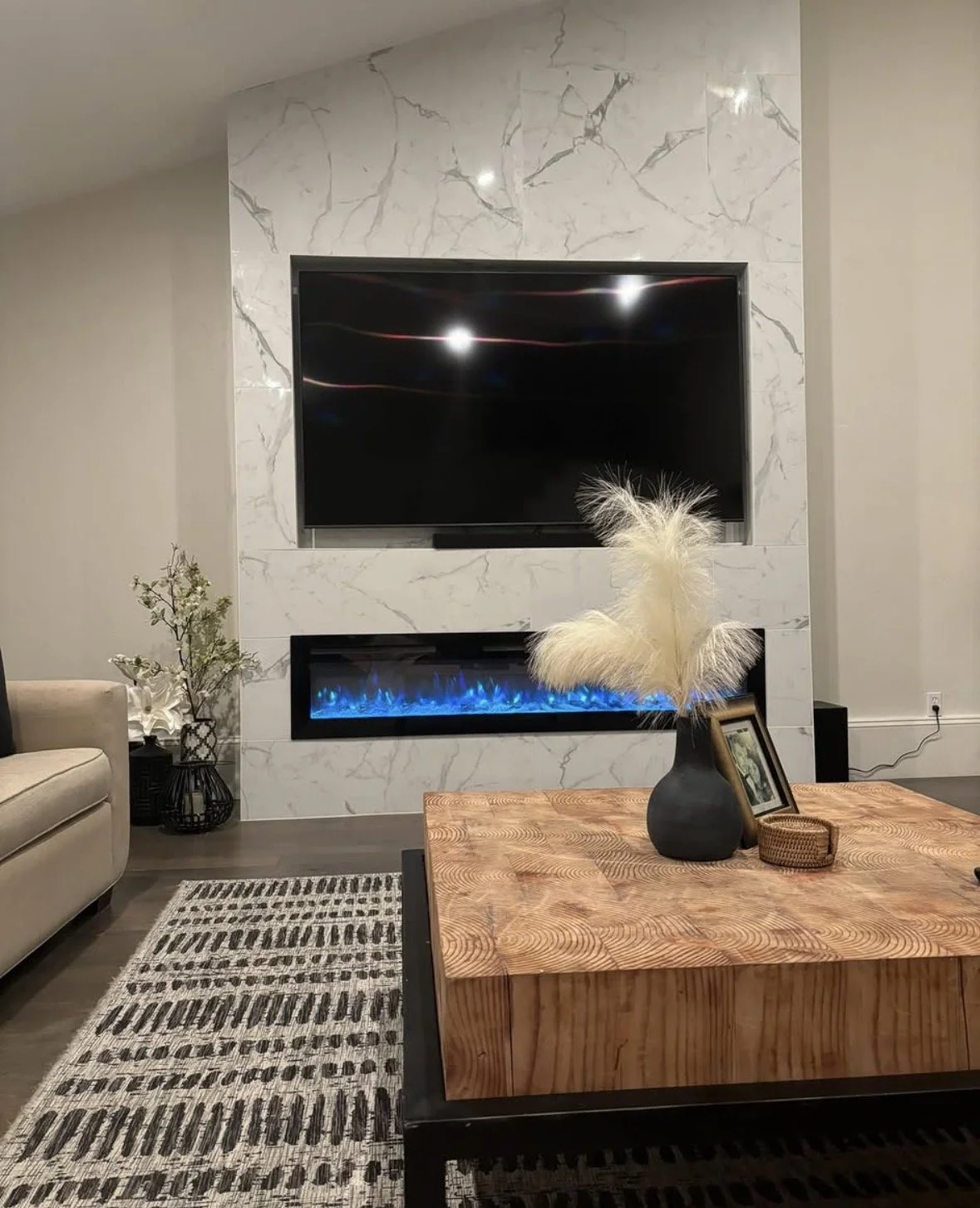 Living room with a mounted flat-screen TV above a modern white marble fireplace with a blue flame gas feature. A wooden coffee table with decorative items, including a black vase with pampas grass and framed photos, is in the foreground. To the left,