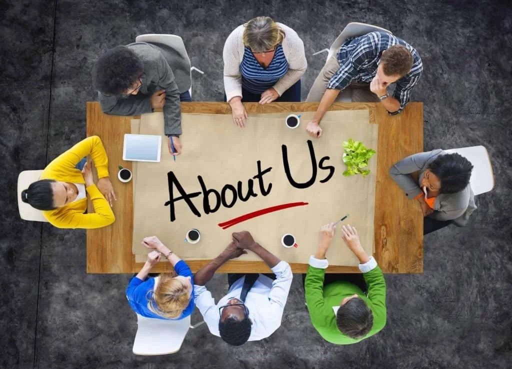 A top-down view of six diverse people sitting around a rectangular wooden table with a beige paper and the words 'About Us' written on it in black marker, several cups of coffee, an iPad, and a small green plant.