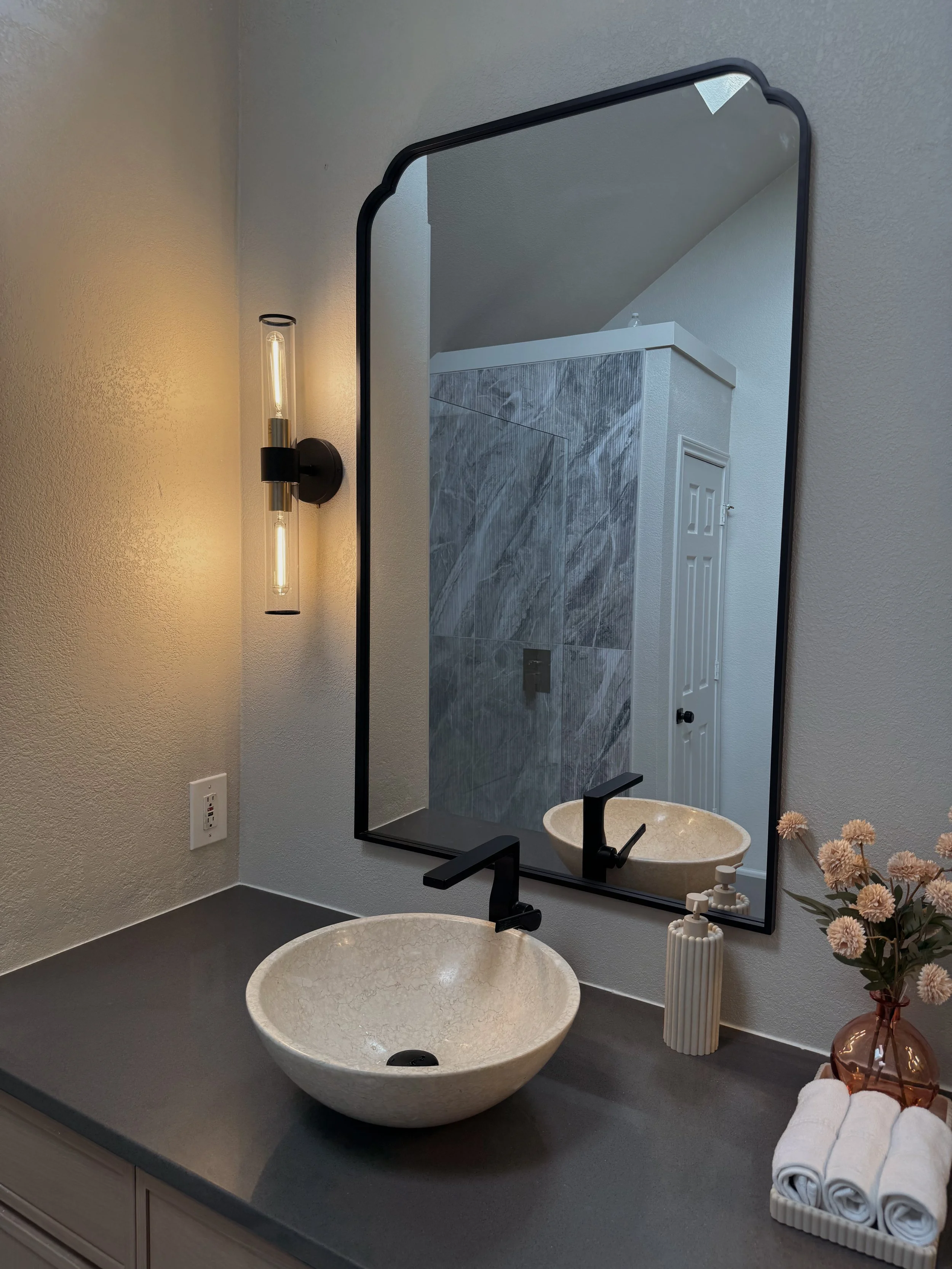 A bathroom vanity with a large round marble sink, a black faucet, a mirror with a black frame, a wall-mounted light, rolled white towels set in a white tray, and decorative flowers in a pink vase.