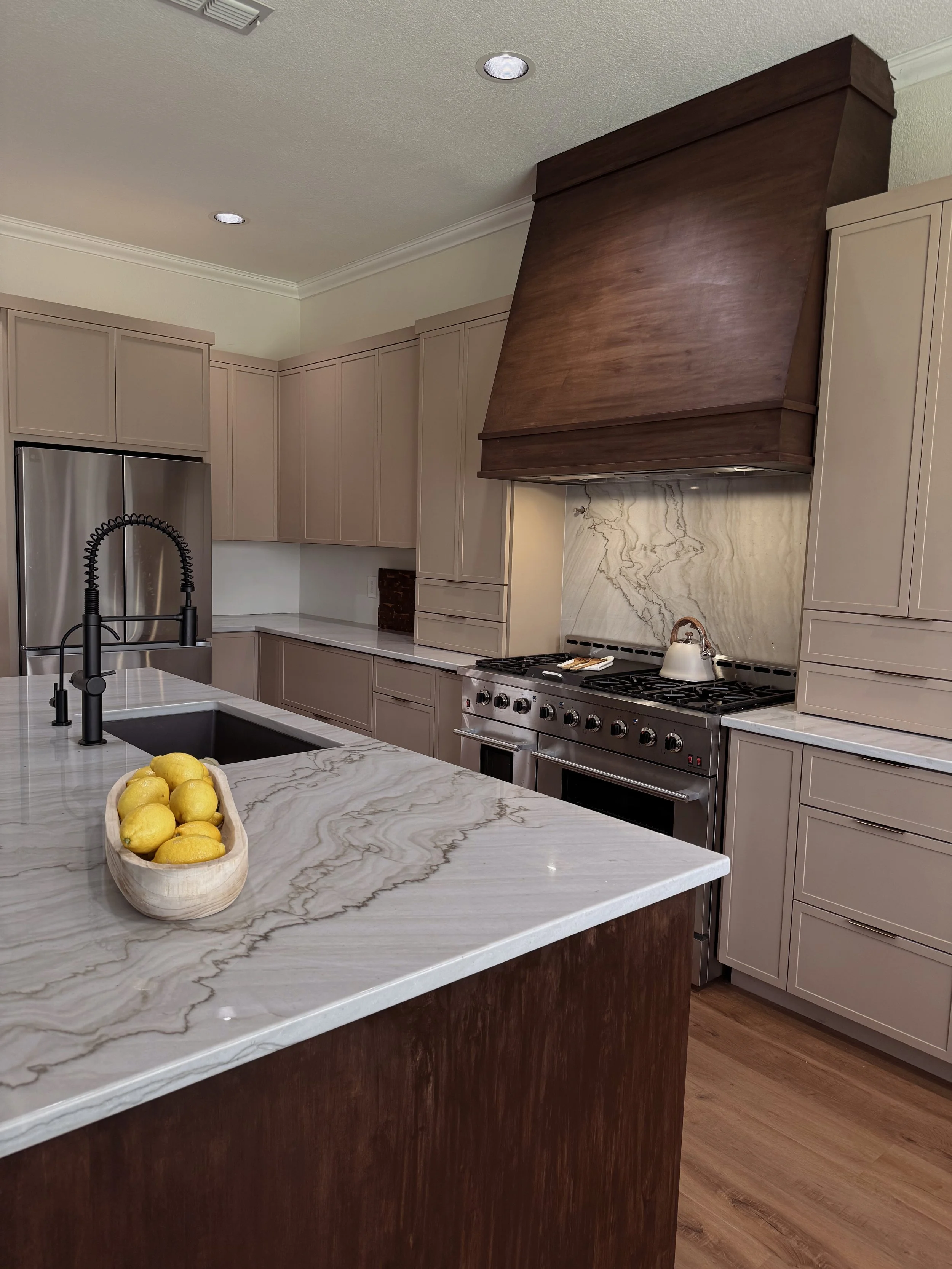 A kitchen with beige cabinets, a large marble-topped island with a bowl of lemons, a black faucet and sink, stainless steel refrigerator, and a stove with a marble backsplash and a wooden range hood.