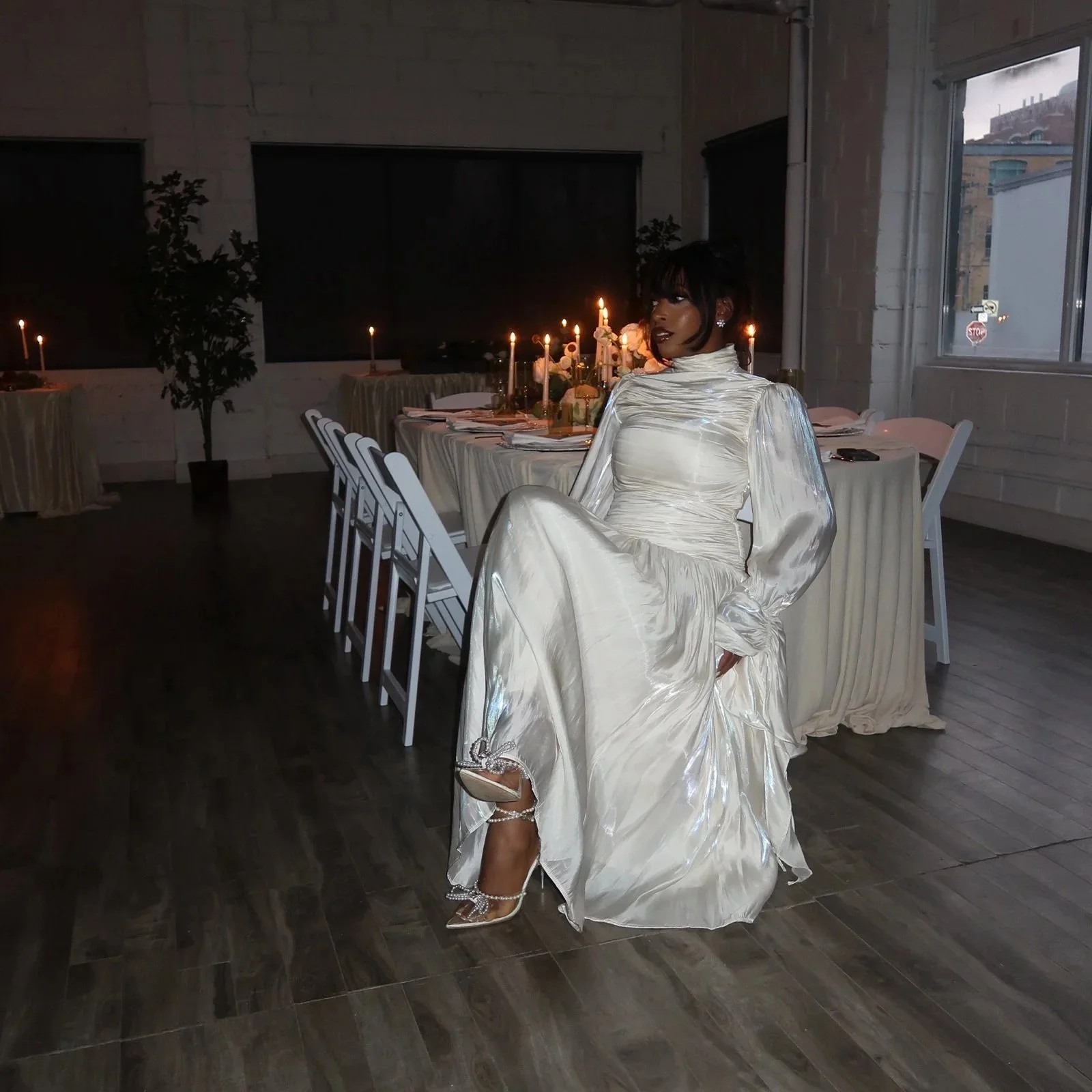 A woman in a shiny, off-white dress with puffed sleeves and high heels sits on a chair in a dimly lit room with decorated tables and candles.