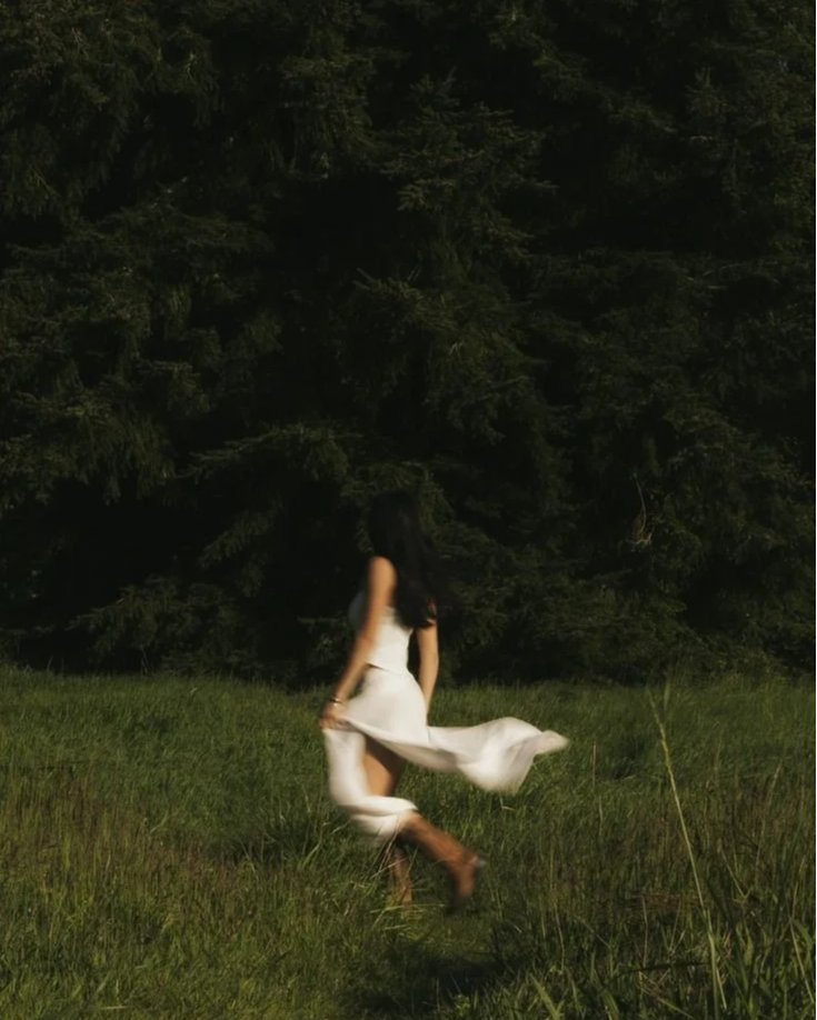 A woman with long dark hair and wearing a white dress is walking through a grassy field with trees in the background.