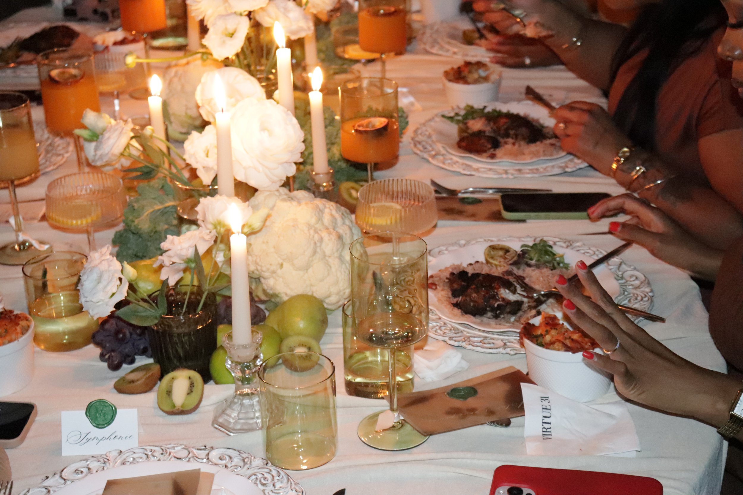 A table decorated with white flowers, candles, and fruit, set for a meal with several plates of food and glasses of drinks. There are people reaching for their plates.