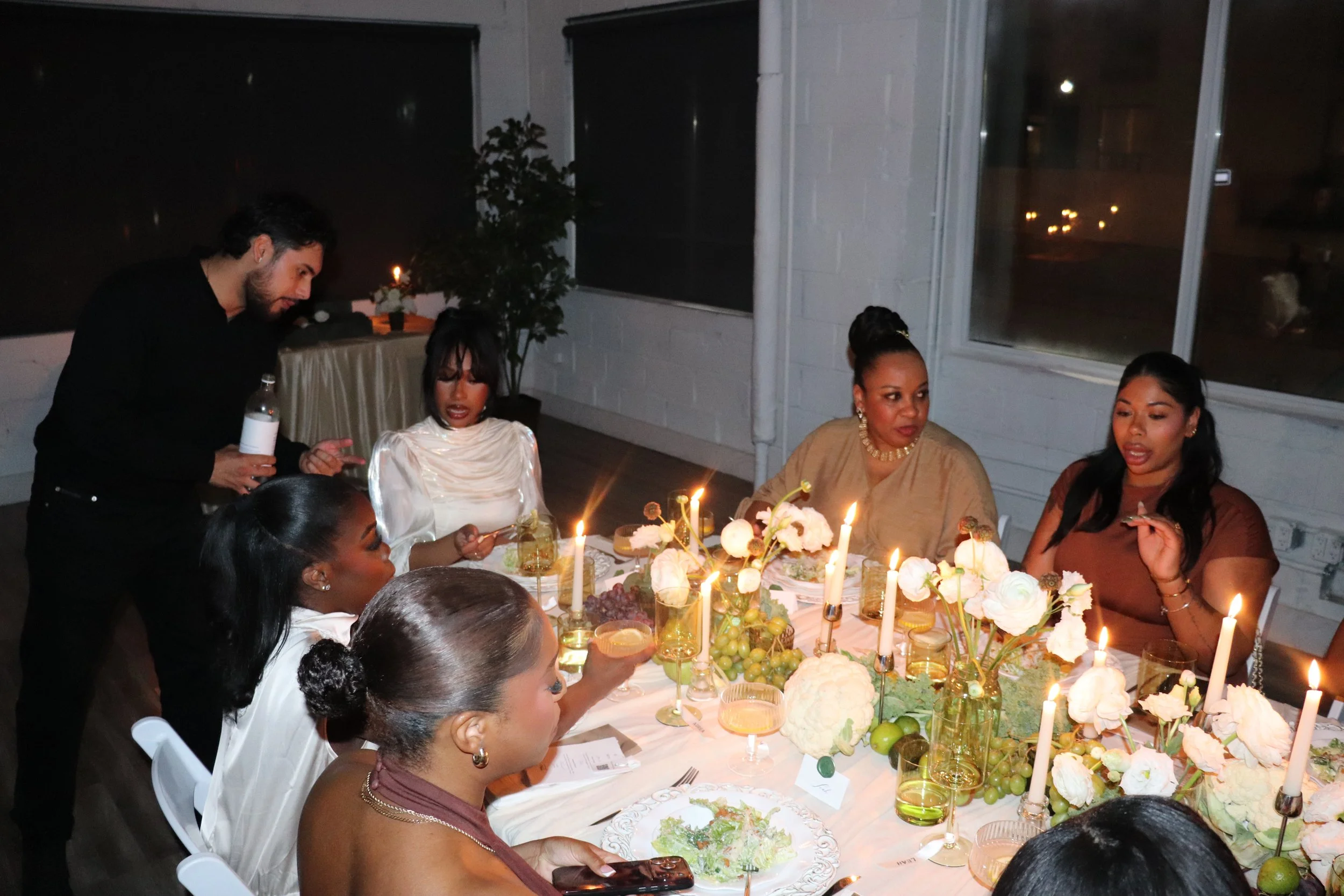 A group of people celebrating a birthday dinner around a decorated table with flowers and candles at night, indoors, with windows in the background.