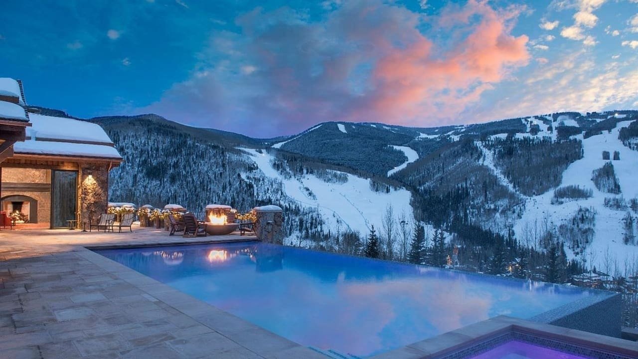 A luxurious mountain retreat with an infinity pool overlooking snow-covered ski slopes and forested mountains at sunset or dusk.