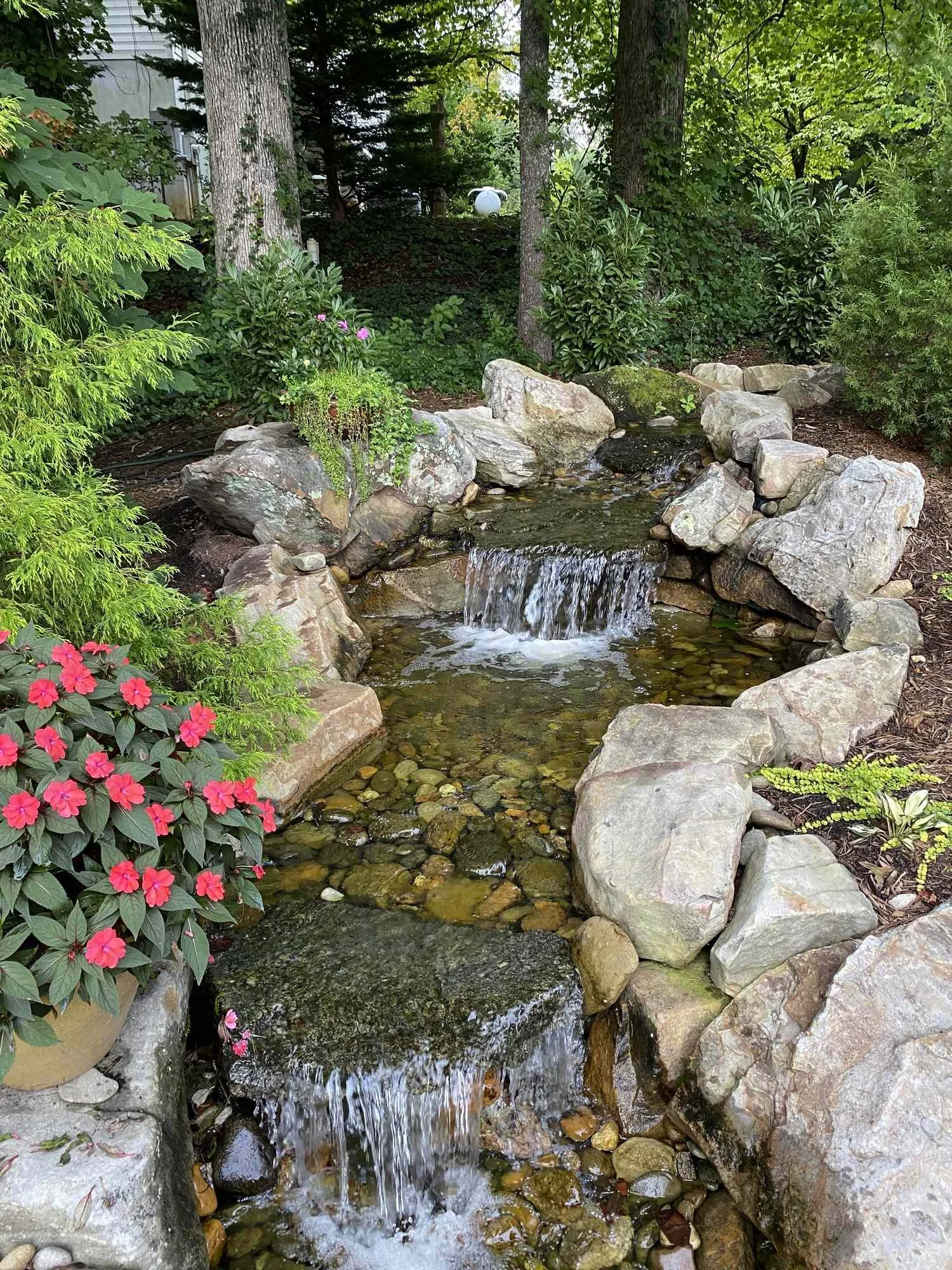 Small waterfall flowing over rocks, surrounded by lush green plants and trees, with pink flowers in a pot on the left.