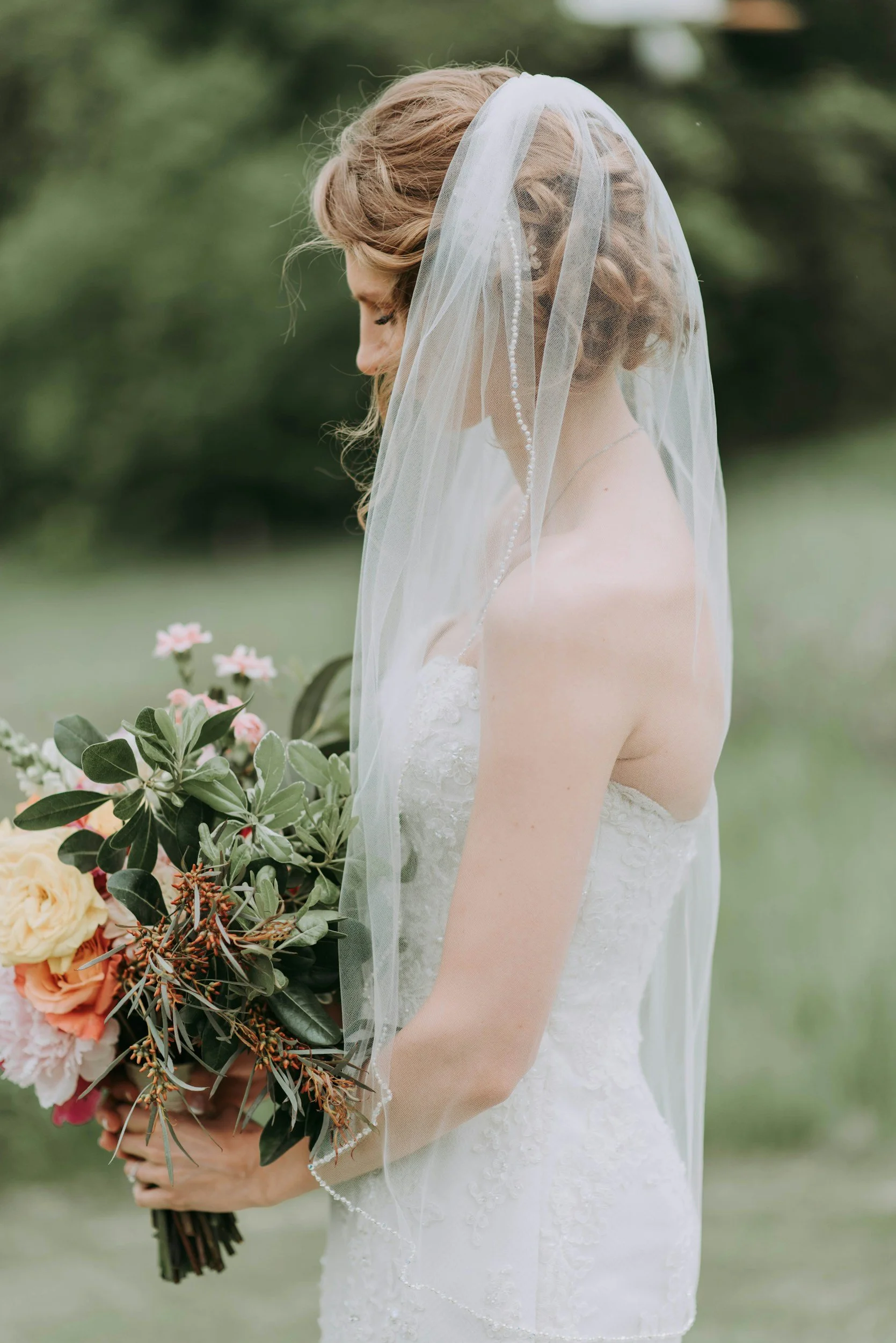 A bride with curly hair in a white wedding dress holding a bouquet of pink and white flowers and green foliage, outdoors in a natural setting with blurred greenery in the background.