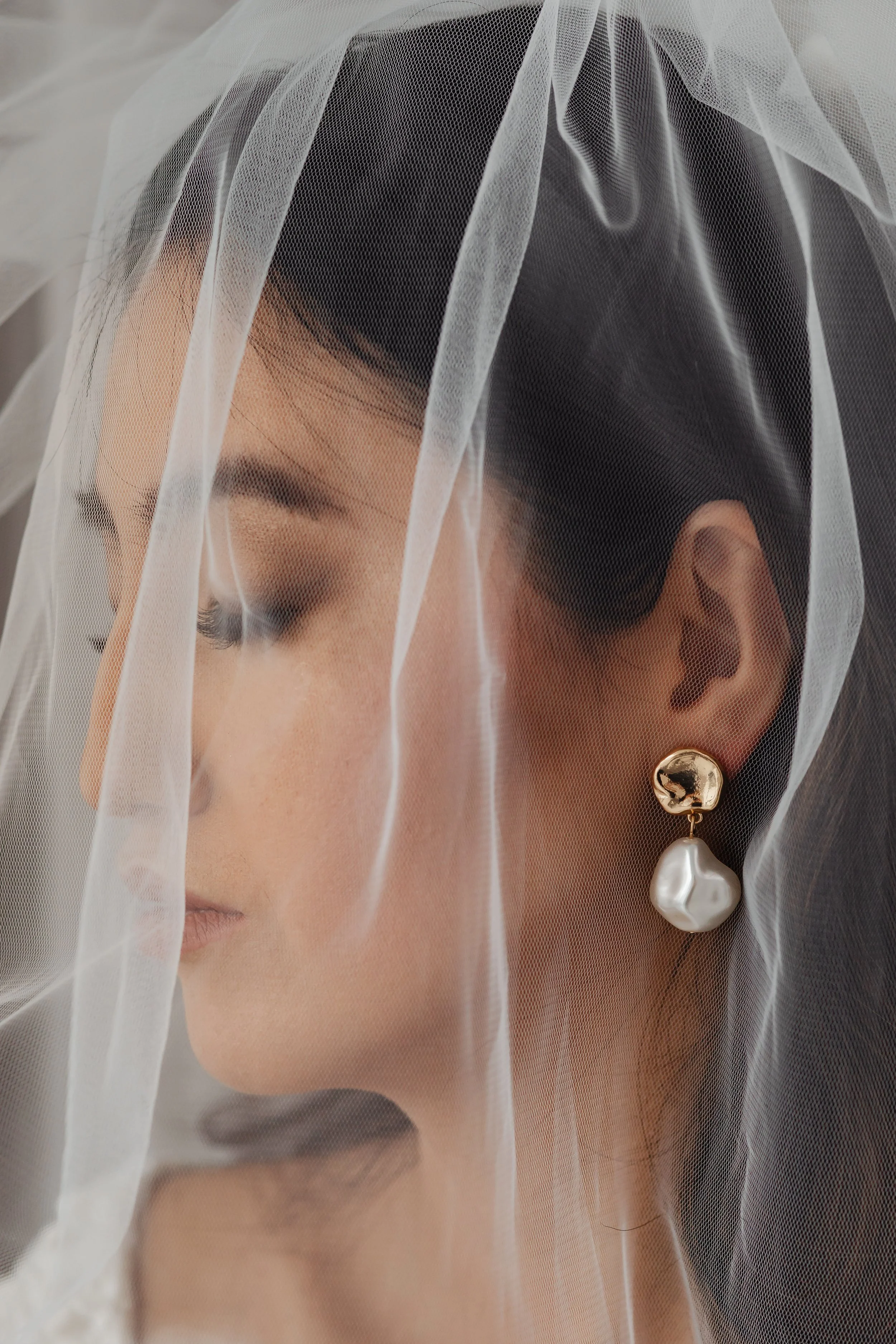 A close-up of a woman with dark hair, wearing a veil and gold earrings with pearl drops.