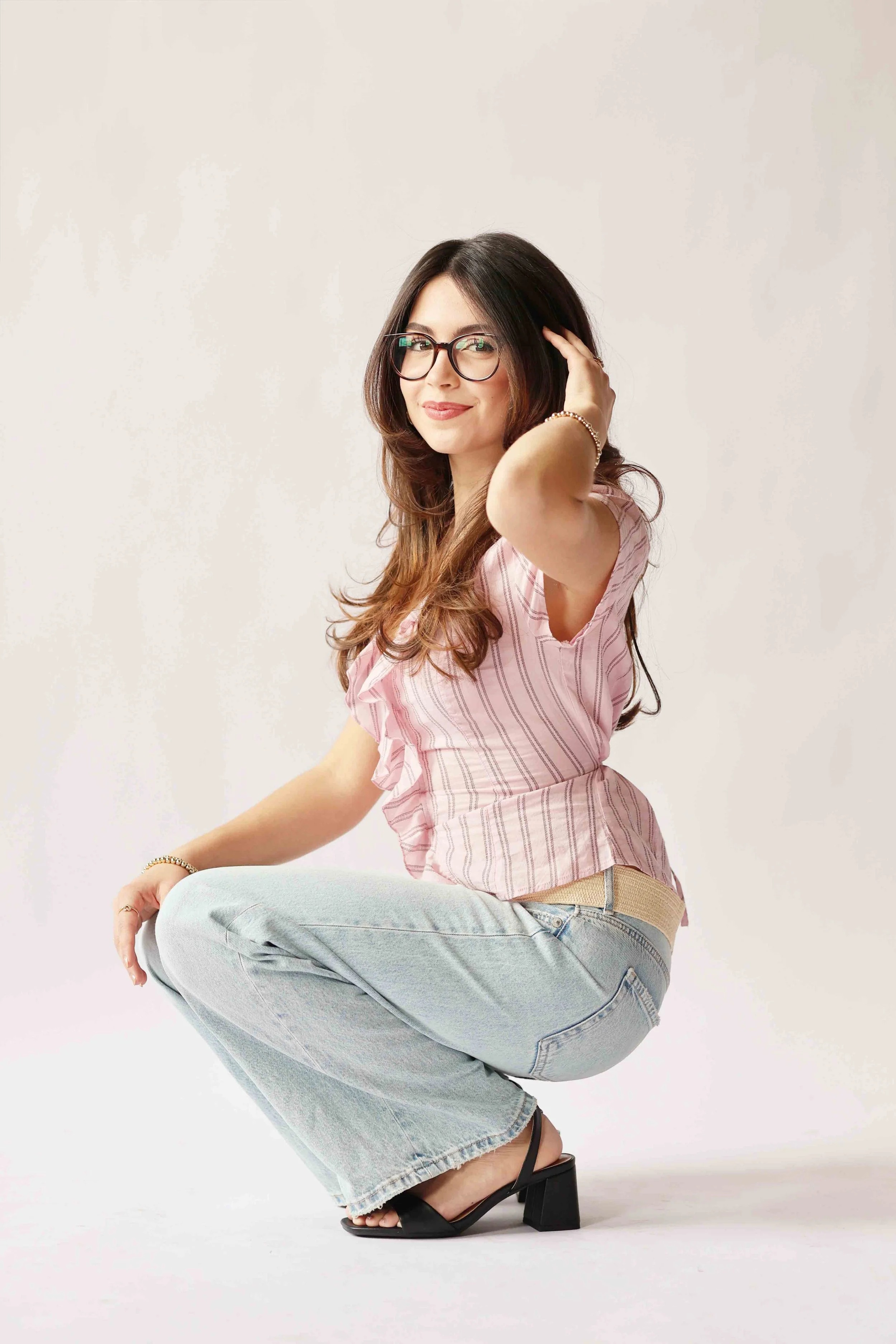 A woman with long brown hair, wearing glasses, a pink striped top, light jeans, and black heeled sandals, smiling and touching her hair, posing in a squatting position against a plain white background.