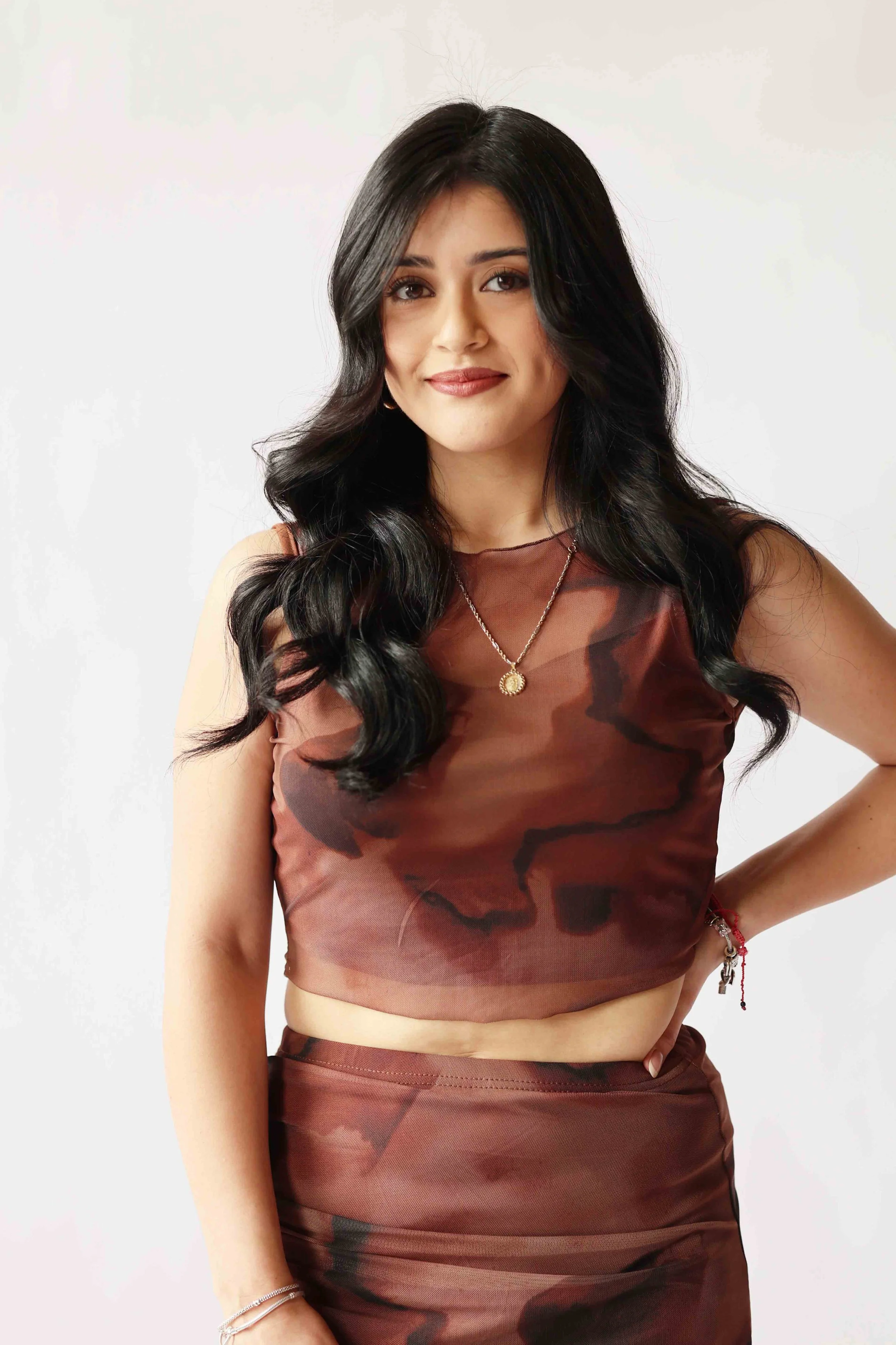 A woman with long black hair styled in loose waves, wearing a sleeveless brown camouflage-patterned top, a gold necklace, and multiple bracelets, standing against a plain white background.