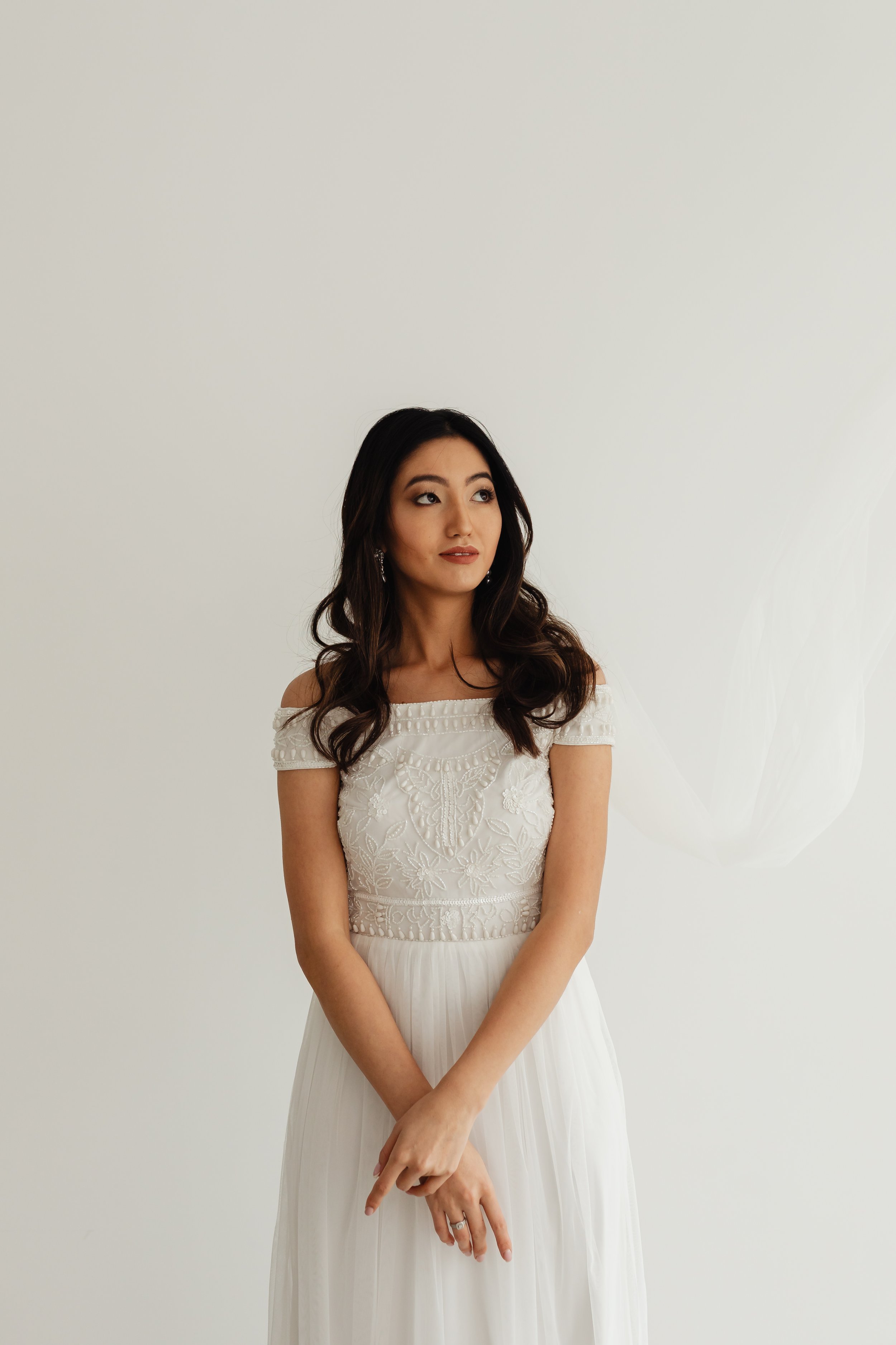 A woman wearing a white off-the-shoulder dress with embroidered details, standing against a plain white wall, with her hands politely crossed in front of her.