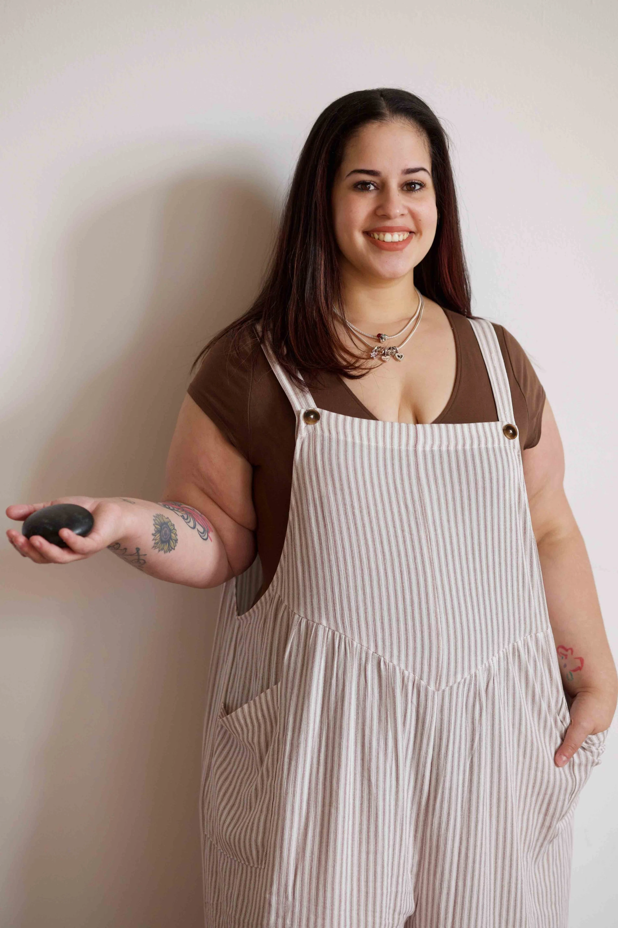 A woman with dark brown hair wearing a brown shirt and beige striped apron stands against a plain white wall, smiling, and holding a remote control in her right hand, with tattoos on her left forearm.