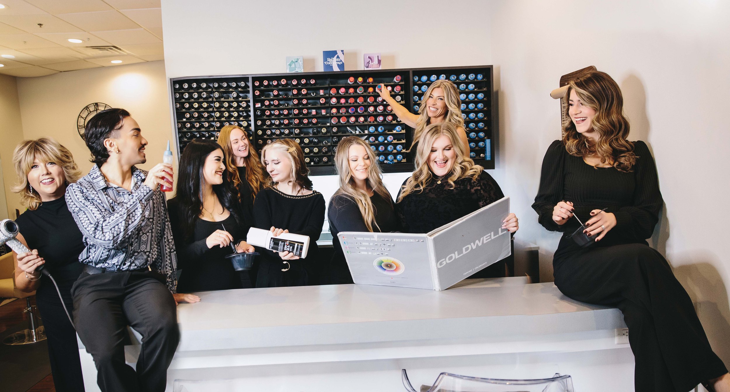 Group of women in a hair salon, some holding hair styling tools and products, smiling and engaging with each other.