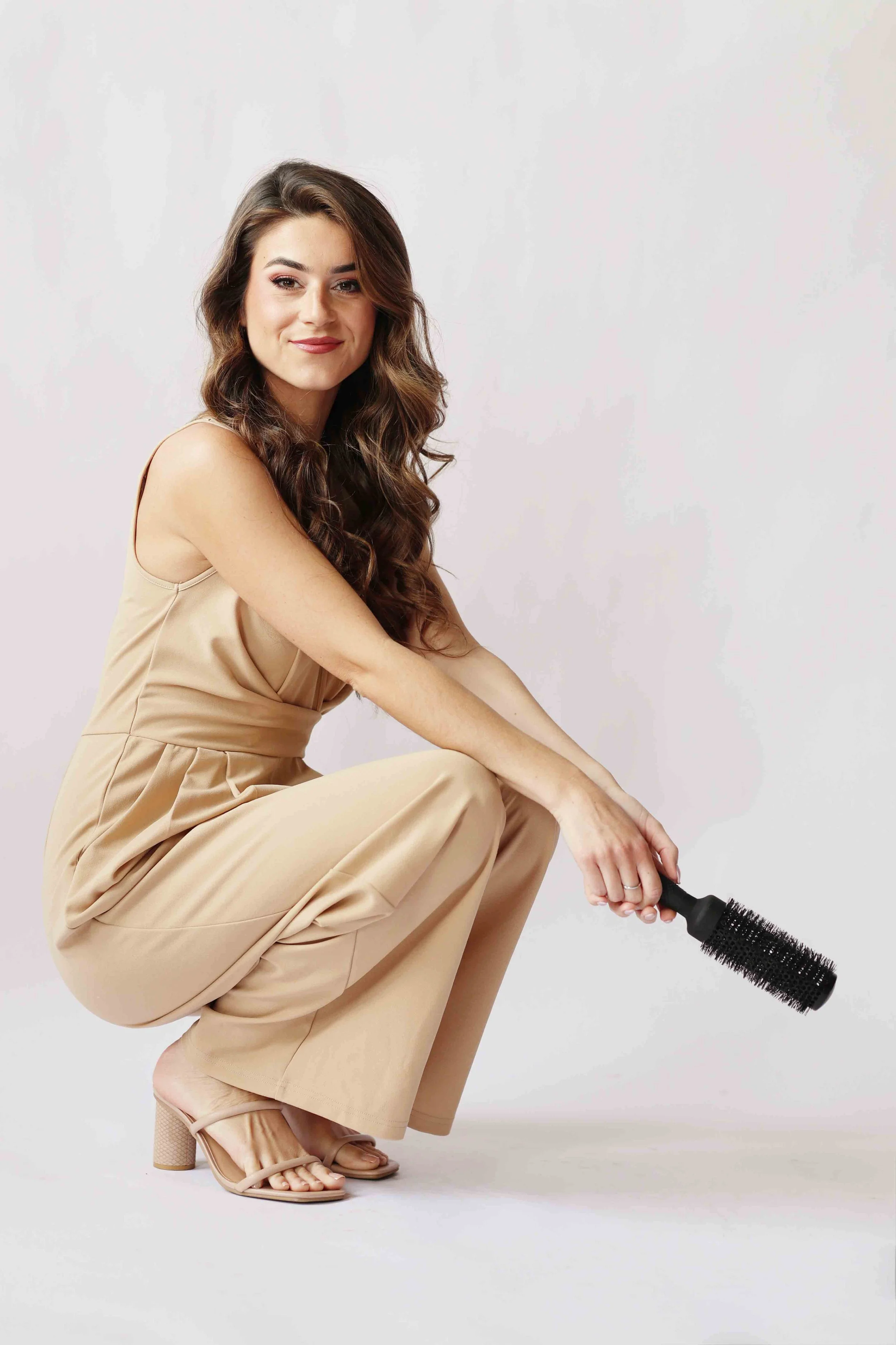 A woman with long, wavy brown hair, wearing a beige sleeveless dress, is squatting while holding a round hairbrush with a handle and bristles, against a plain light background.