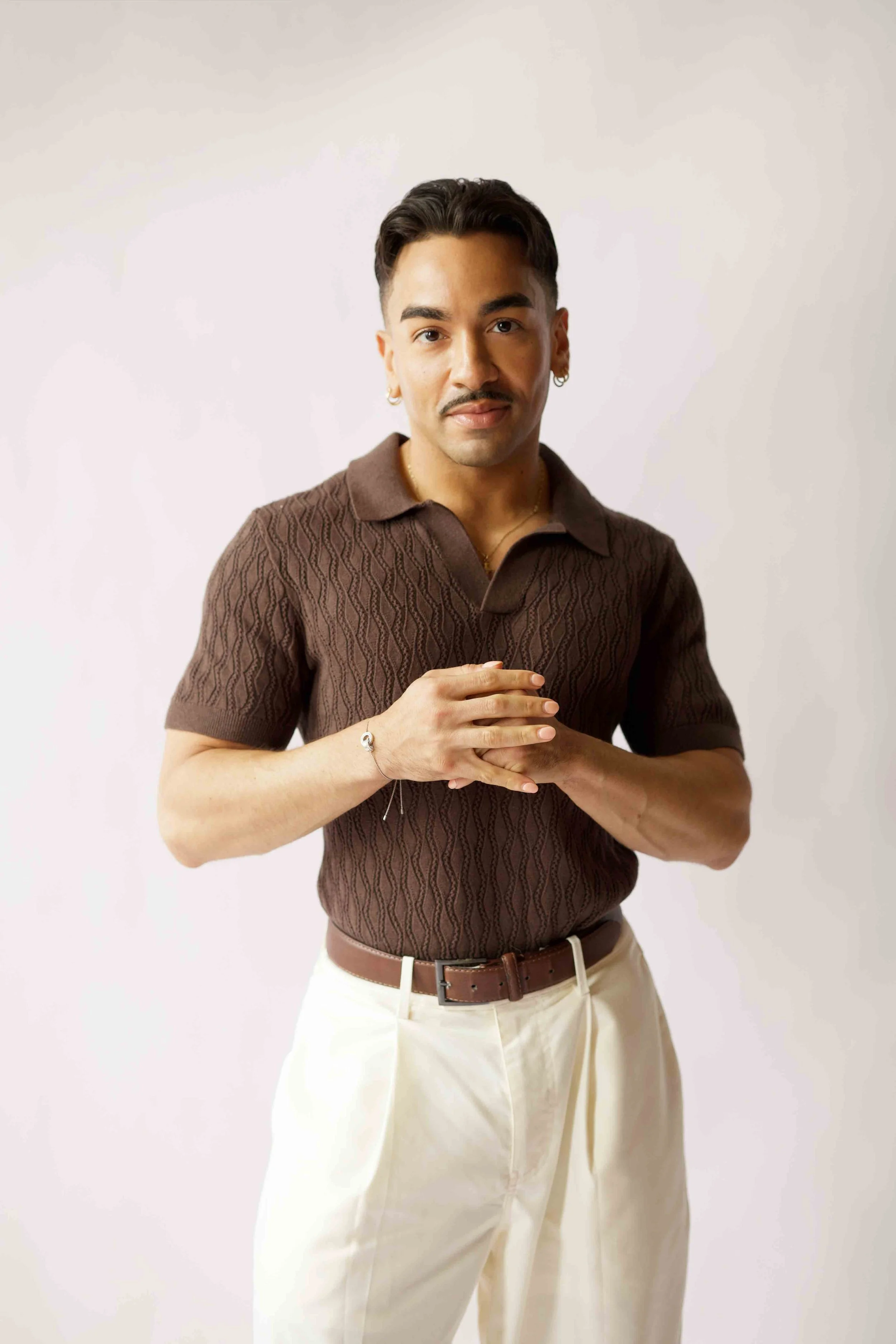 A person with medium skin tone, dark hair, and a mustache stands against a plain light background. They are wearing a brown, patterned short-sleeved polo shirt, beige pants, and a brown belt. They have earrings and a bracelet, and their hands are clasped together at chest level.