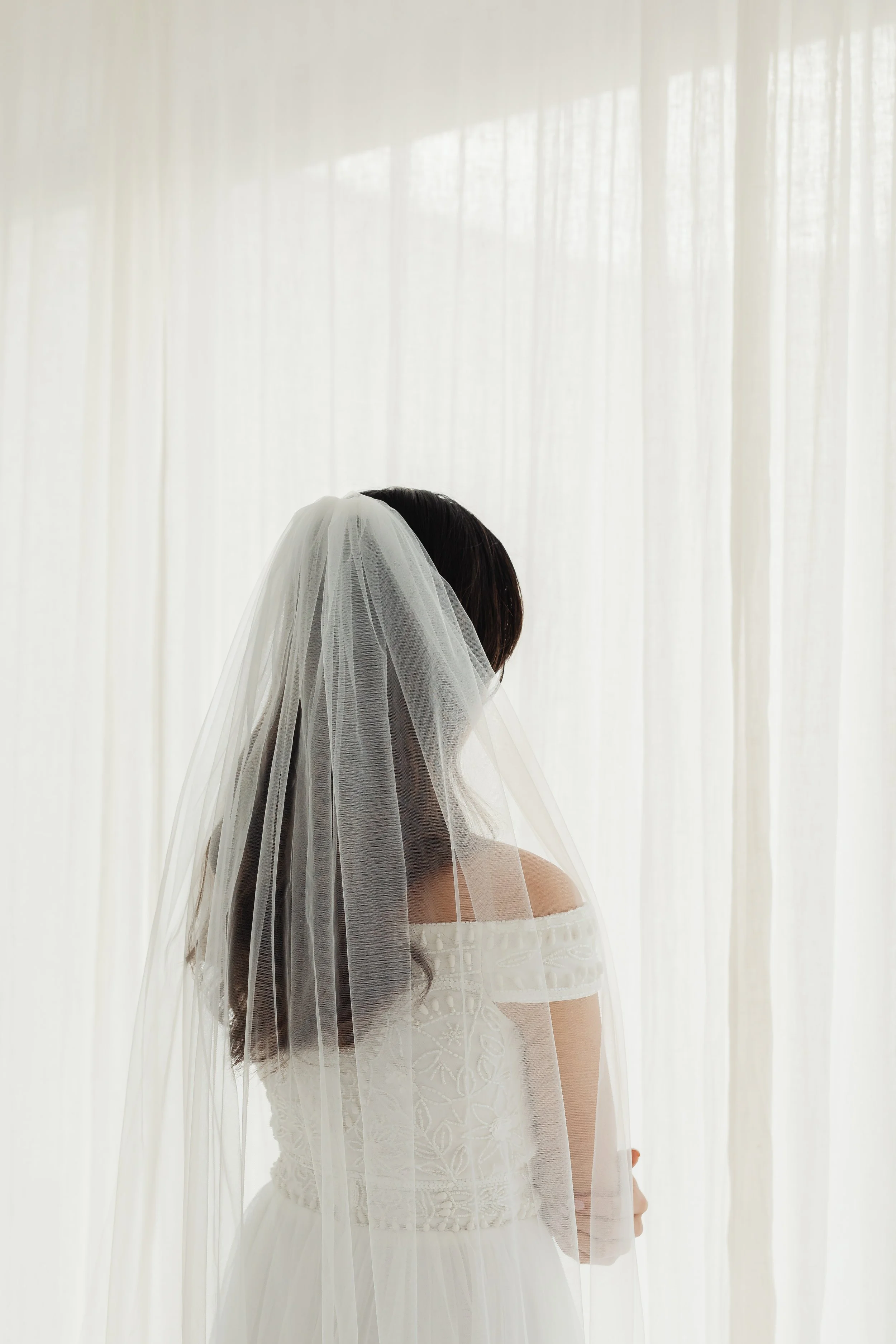 A bride in an off-shoulder white dress with lace details, wearing a veil, standing in front of sheer white curtains, facing away from the camera.