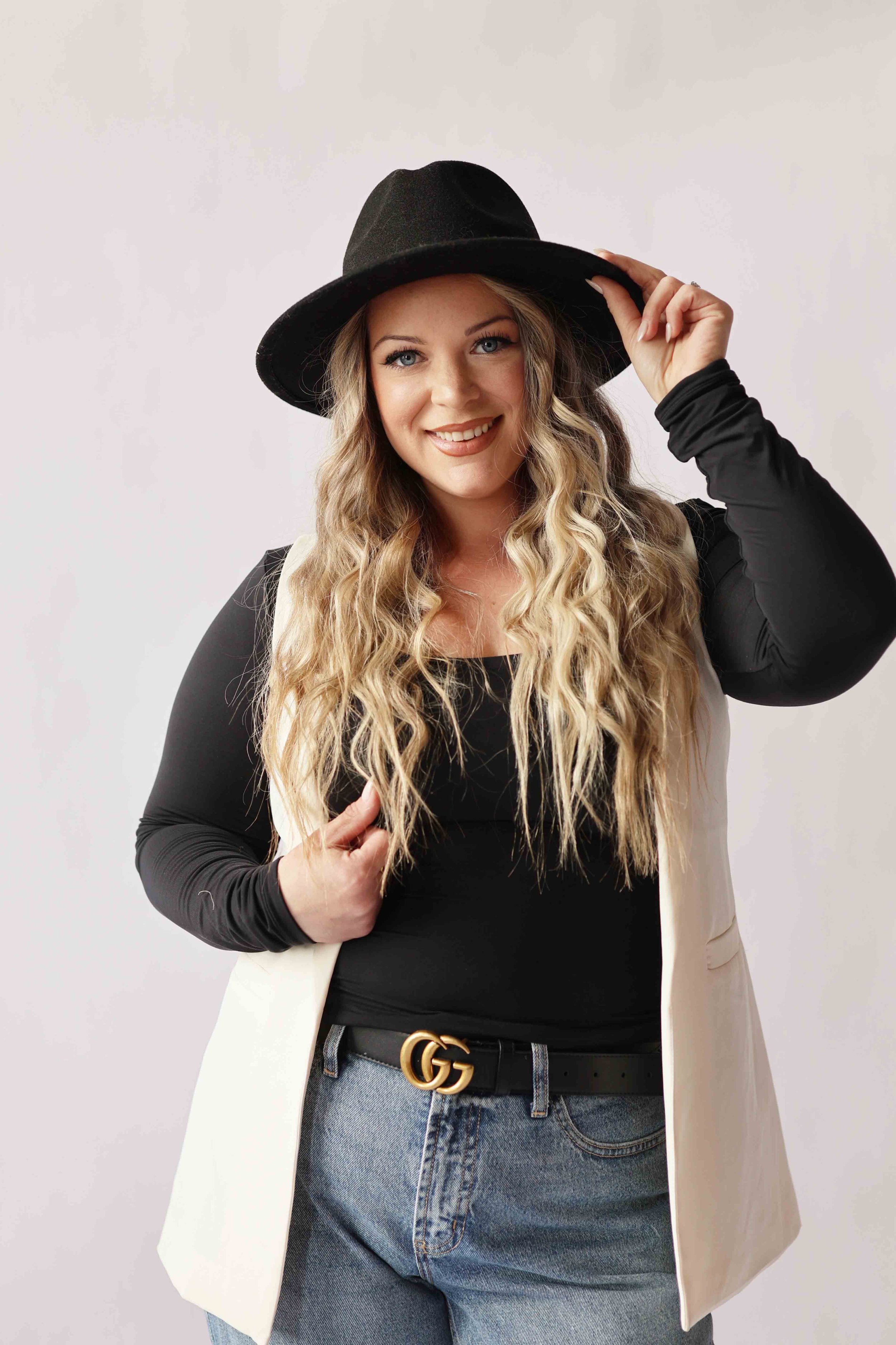 A young woman with long, wavy blonde hair smiling and wearing a black wide-brimmed hat, black long-sleeve top, beige vest, high-waisted jeans, and a Gucci belt, posing against a plain light background.